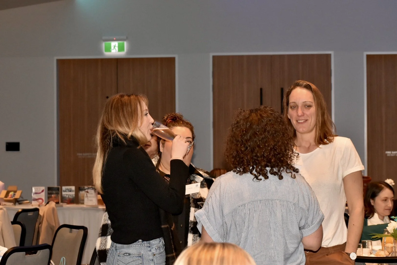 Four women in a social setting, one drinking from a glass, others engaging in conversation at a table in a banquet or conference room.