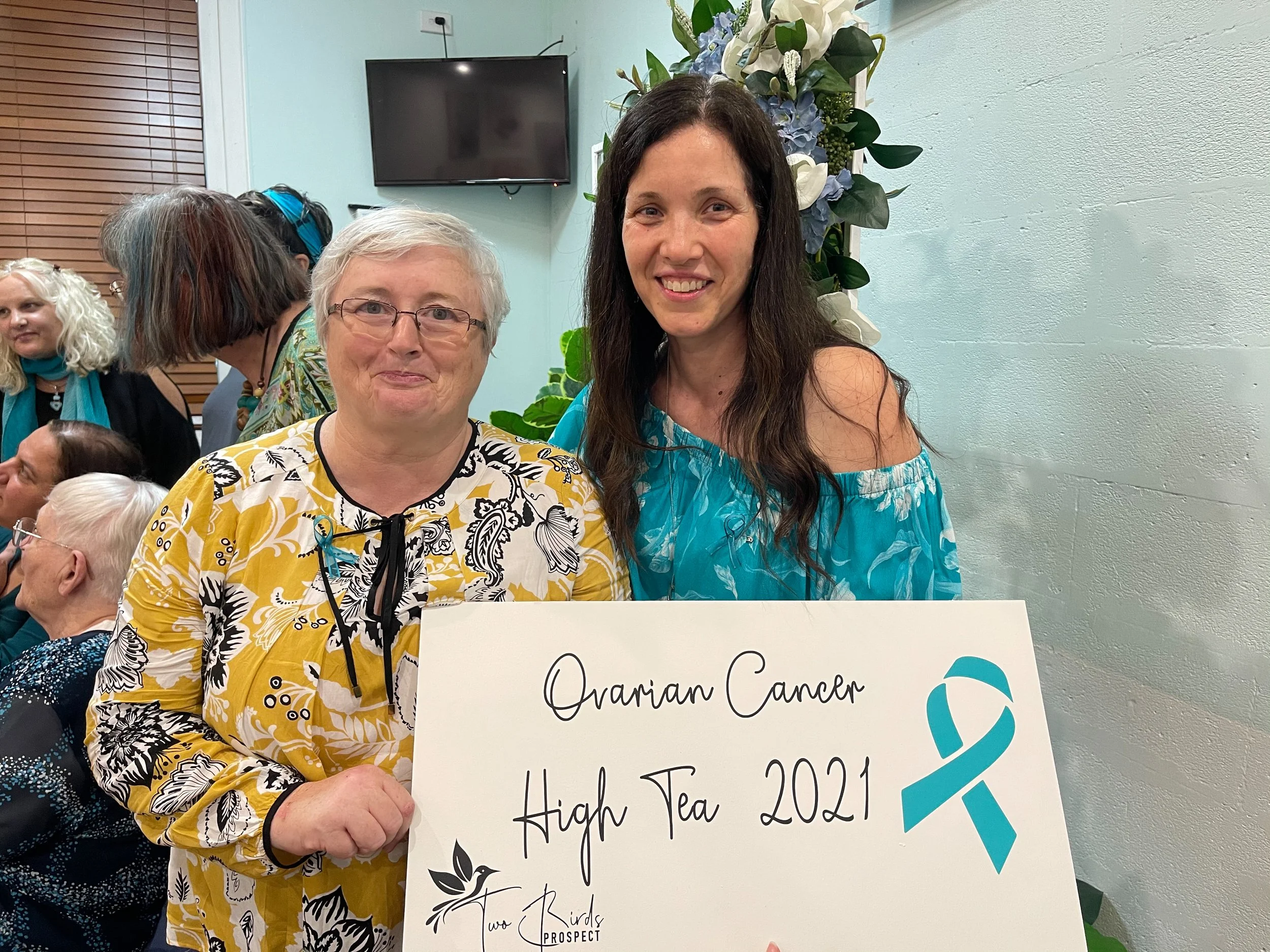 Two women, one with glasses and short gray hair and the other with long dark hair, smiling and holding a sign that reads 'Ovarian Cancer High Tea 2021' with a teal ribbon logo, in a room with other attendees and a large floral arrangement in the back