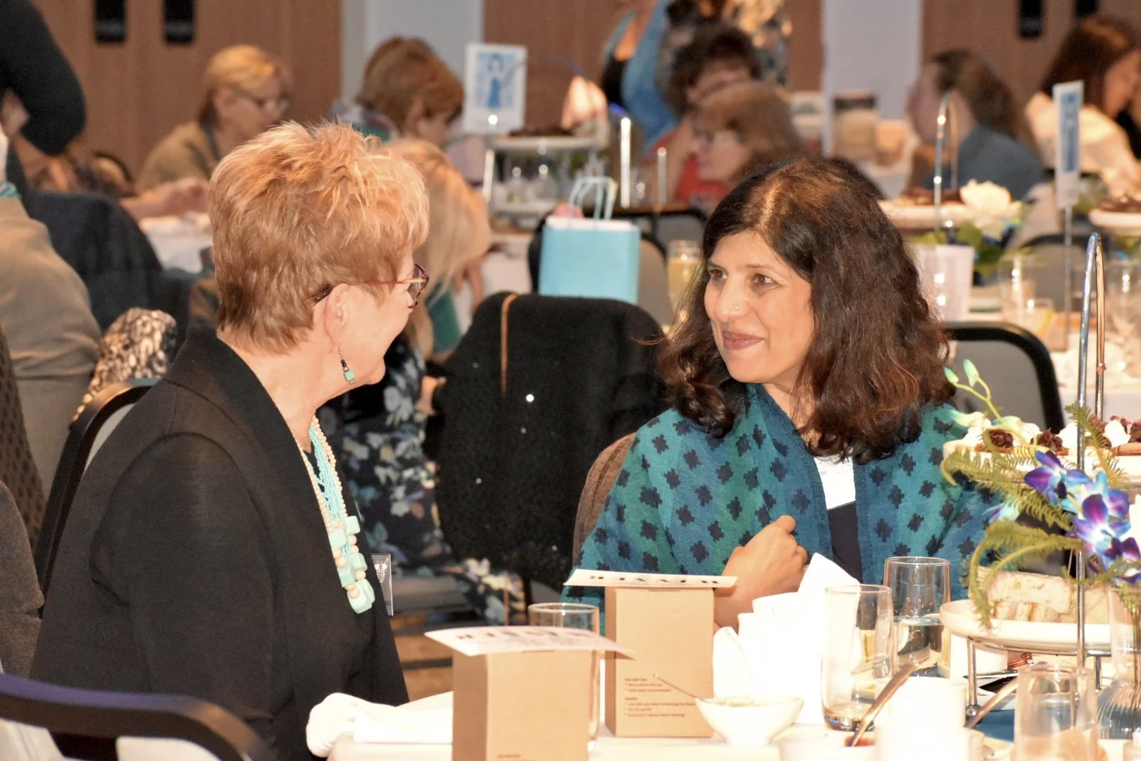 Two women talking at a formal event, seated at a round table with decorated centerpieces and food.