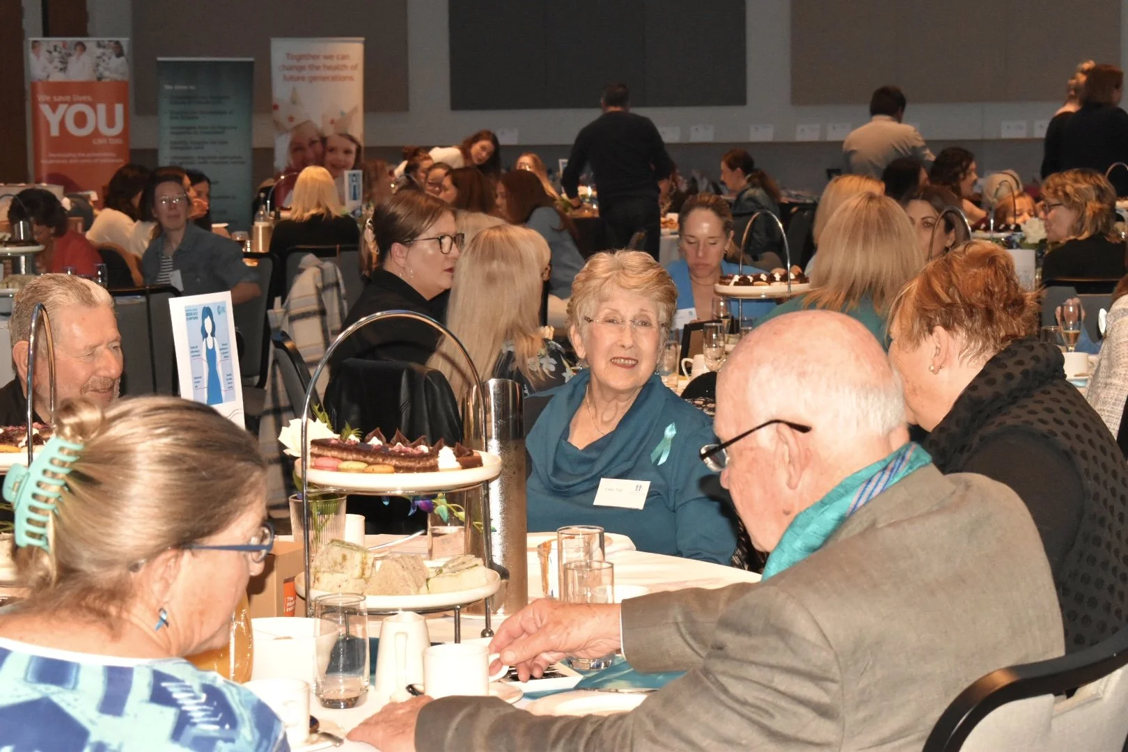 A large group of people seated around conference tables at a formal event, with some engaged in conversation, and a display of cakes and food on the tables.