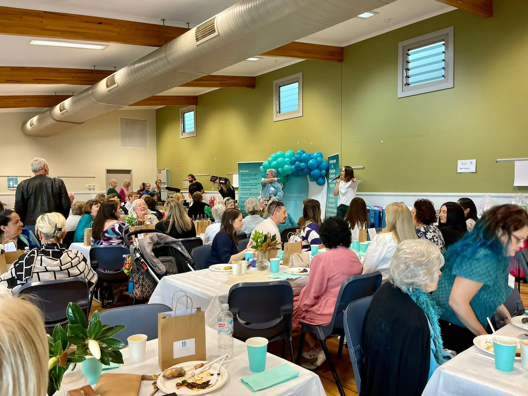 A large gathering of people seated at tables in a community hall, with a stage decorated with blue balloons and a banner related to cancer awareness, indicating a charity event or fundraiser.