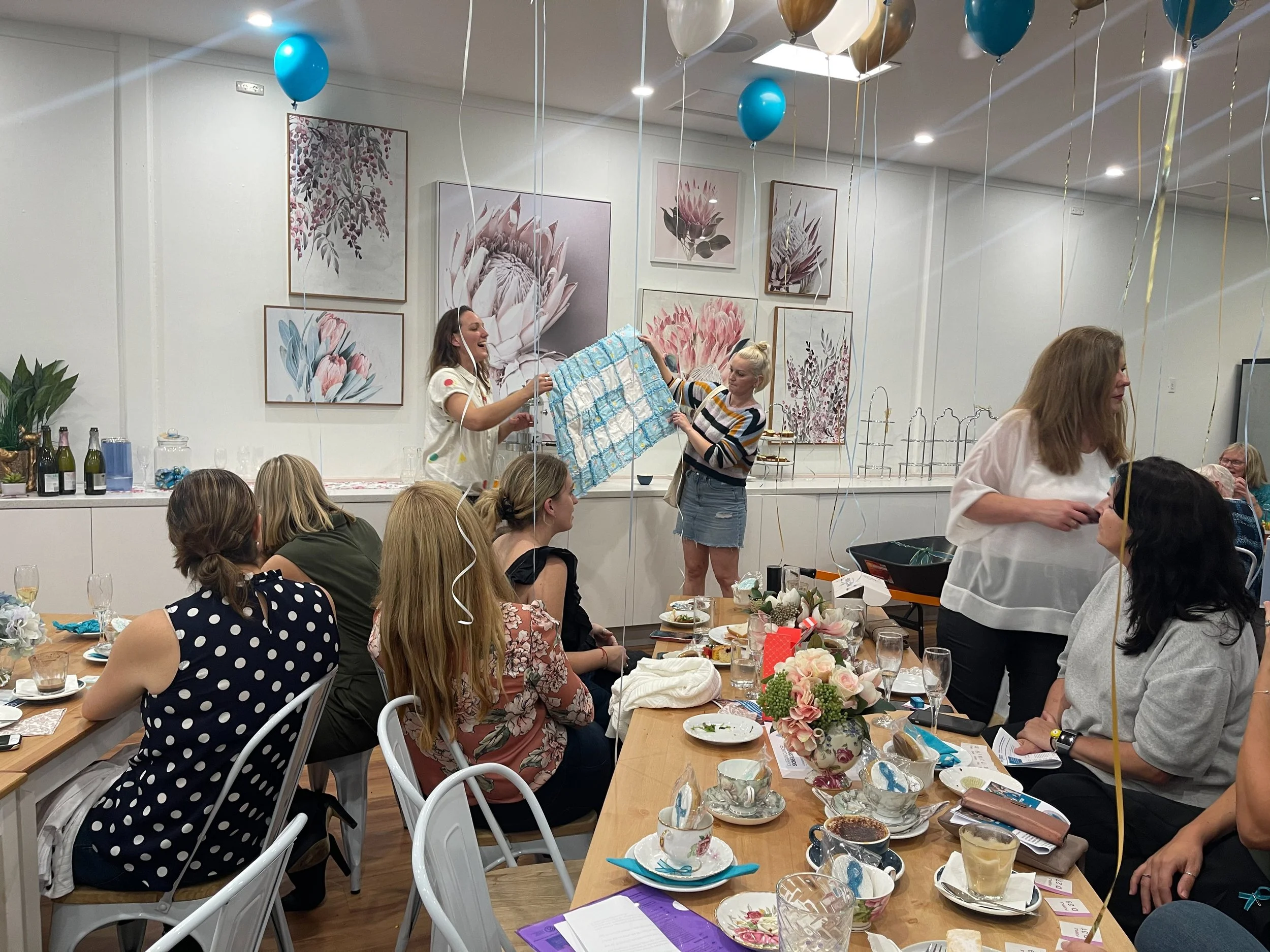People celebrating at a gathering with balloons, food, drinks, and decorations in a decorated room with floral artwork on the walls.