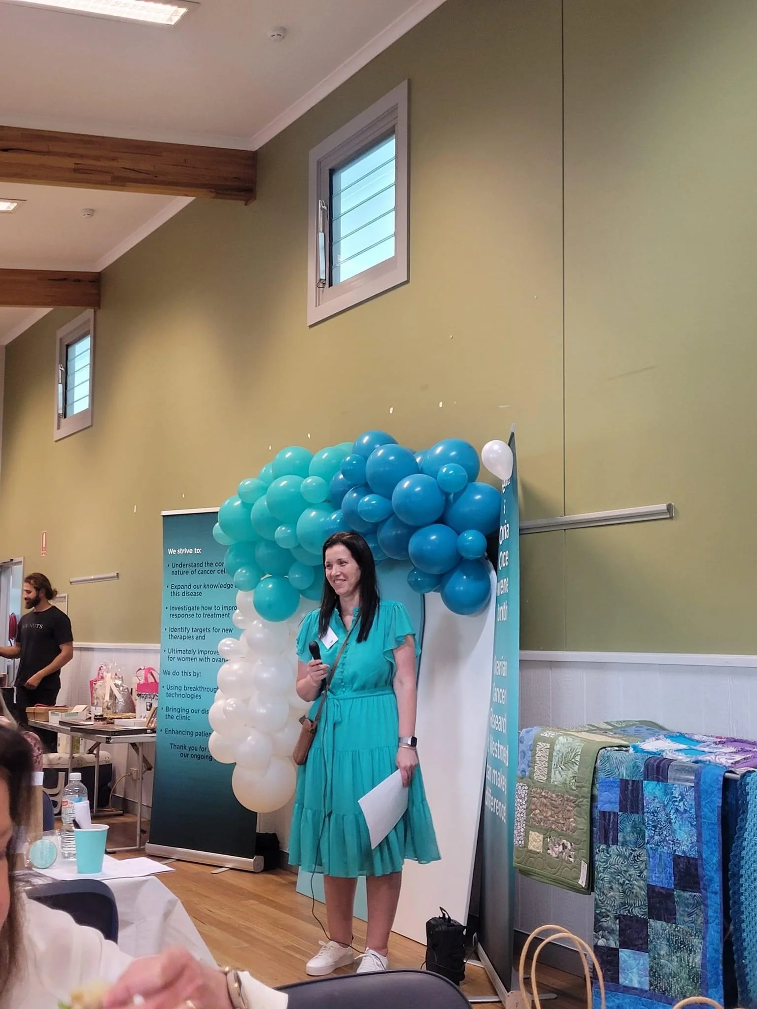 Woman in turquoise dress holding microphone and paper, standing in front of blue and white balloon arch at indoor event.