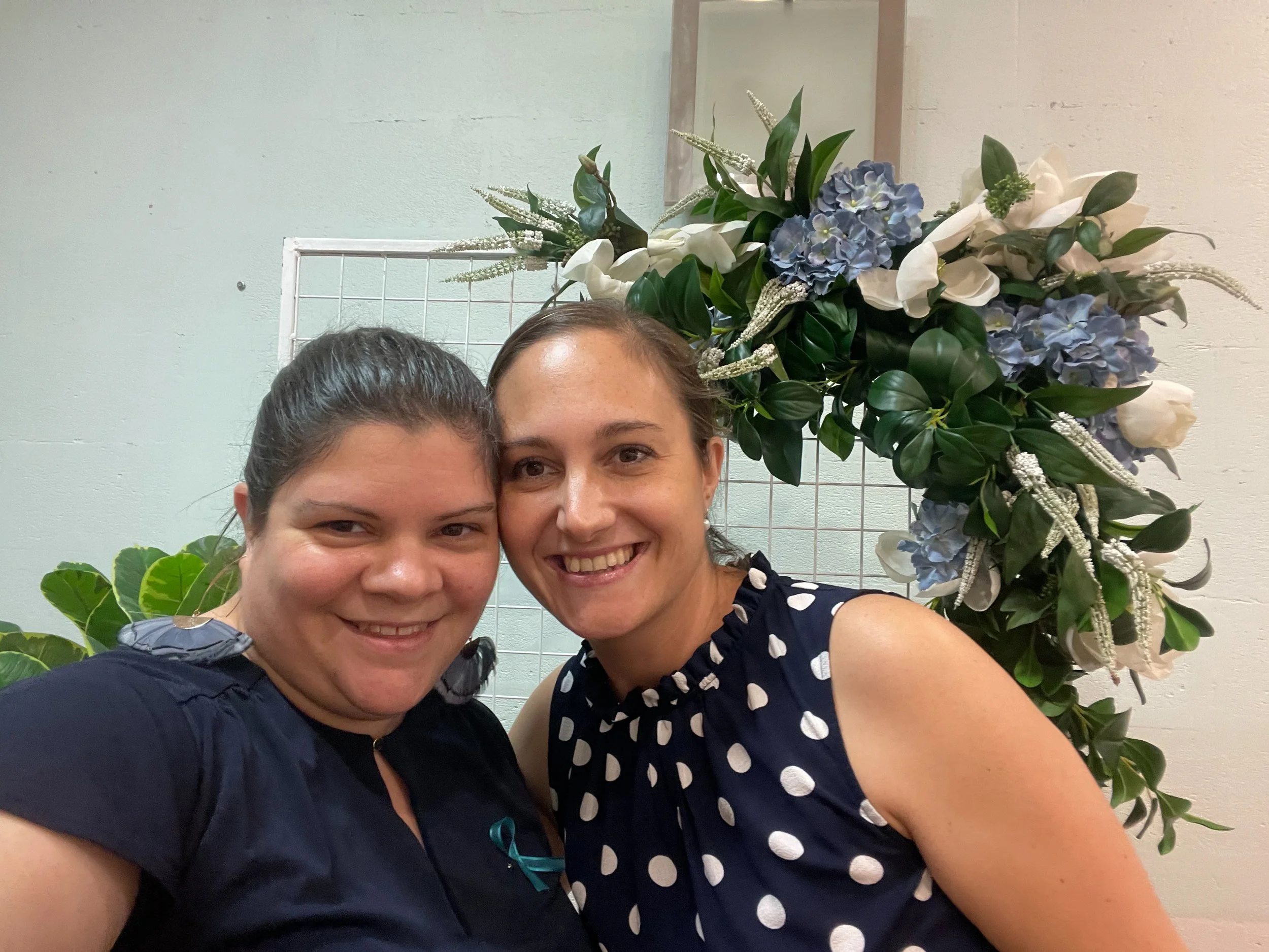 Two women smiling for a photo in front of a floral arrangement with white and light blue flowers and green leaves.