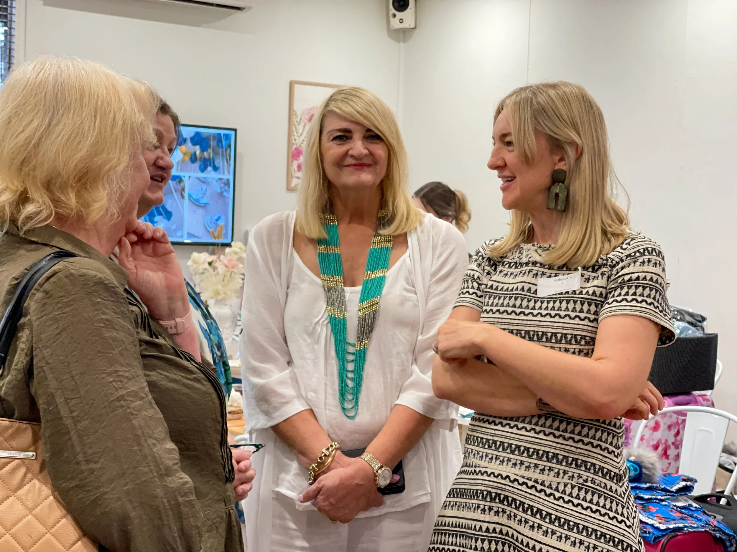 Four women talking at a social gathering, one woman in the middle wearing a white outfit and colorful beaded necklace, another woman on the right in a patterned dress and oversized earrings, the woman on the left with blonde hair wearing a tan jacket