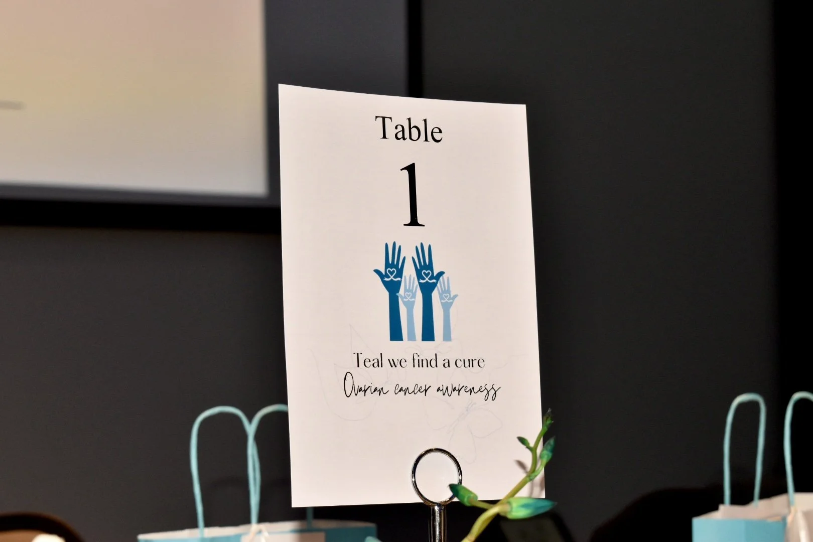 Table sign with the number 1, featuring blue and white hands with hearts, and text about ovarian cancer awareness.