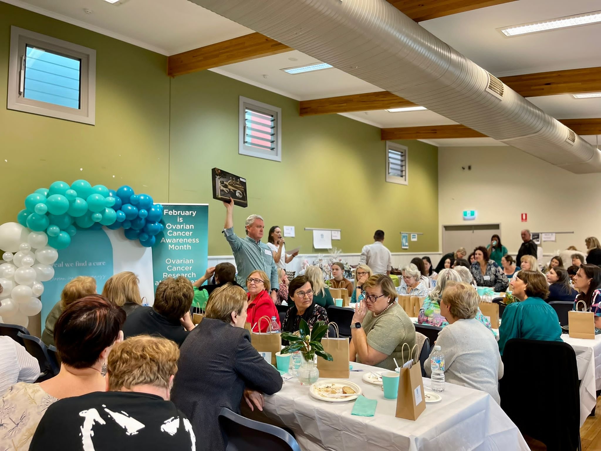 A gathering of people in a community center for Ovarian Cancer Awareness Month, with a speaker holding a plaque, decorated with balloon arch, seated at tables, eating and listening.