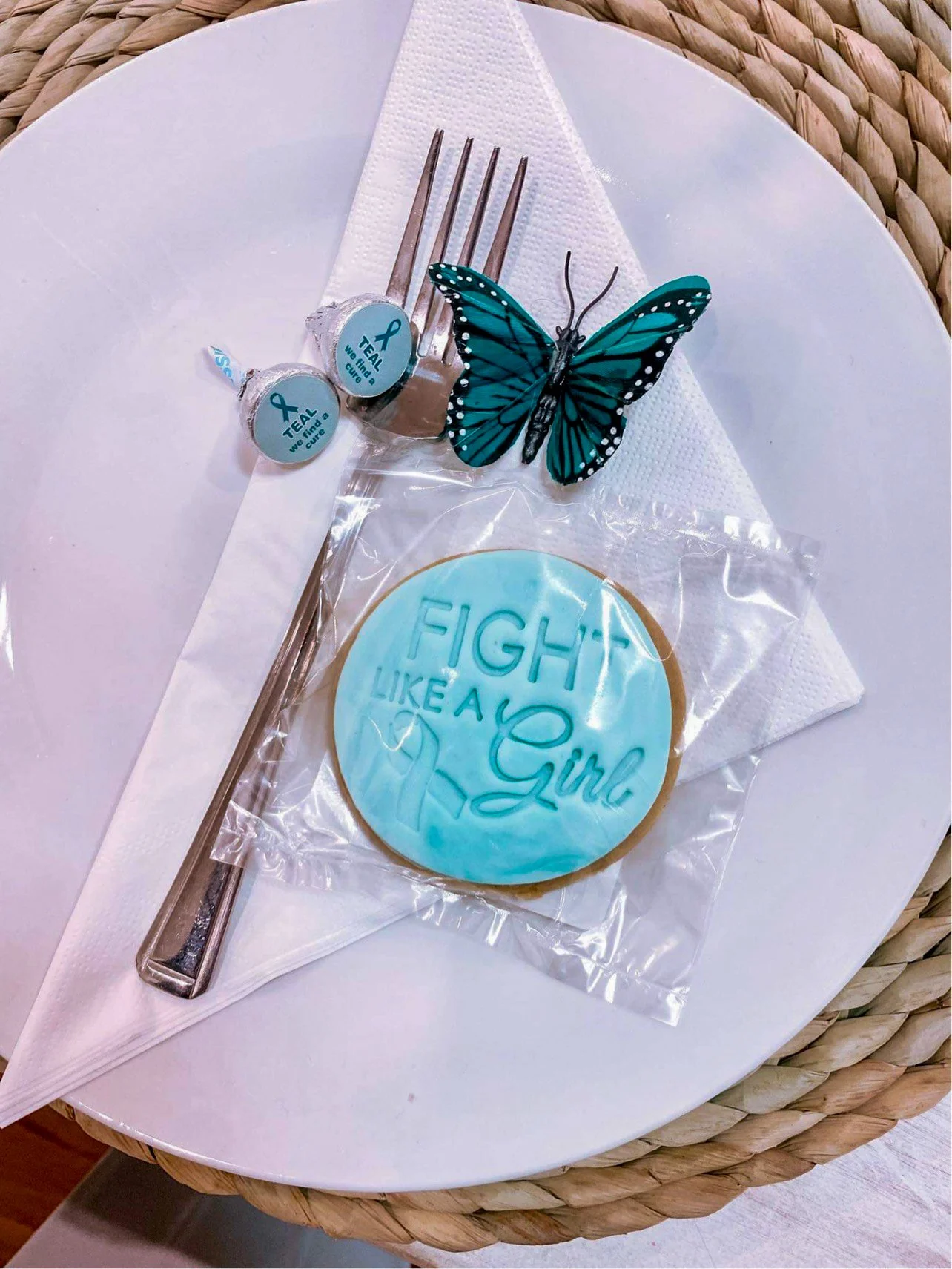 A table setting with a fork, two teal cancer awareness candies, a teal butterfly decoration, and a round cookie with teal icing that reads 'Fight Like a Girl' in script, all on a white plate with a napkin.