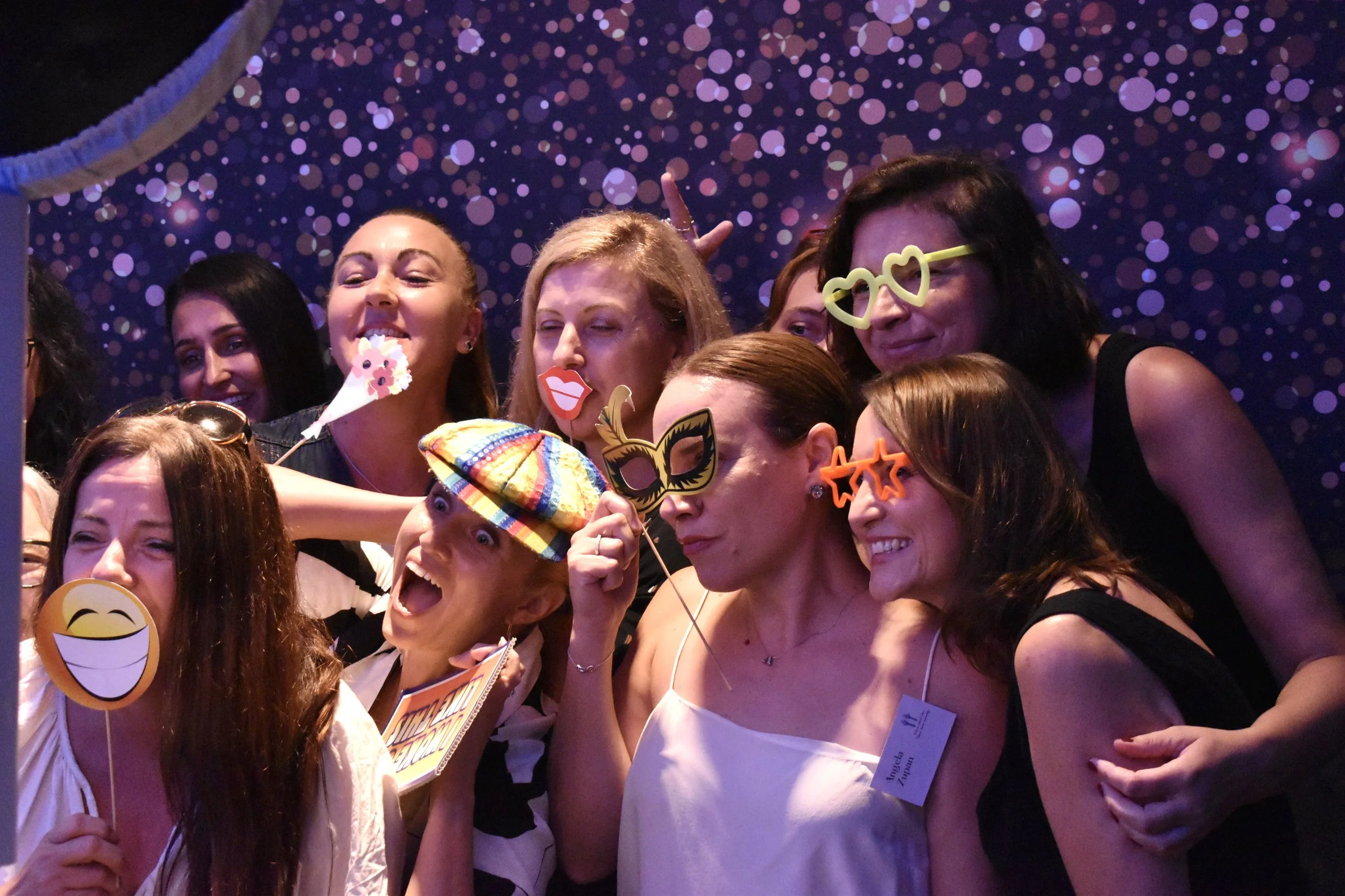 Group of women celebrating at a party, wearing humorous sunglasses and holding photo booth props, with a dark purple backdrop decorated with small pink and purple dots.