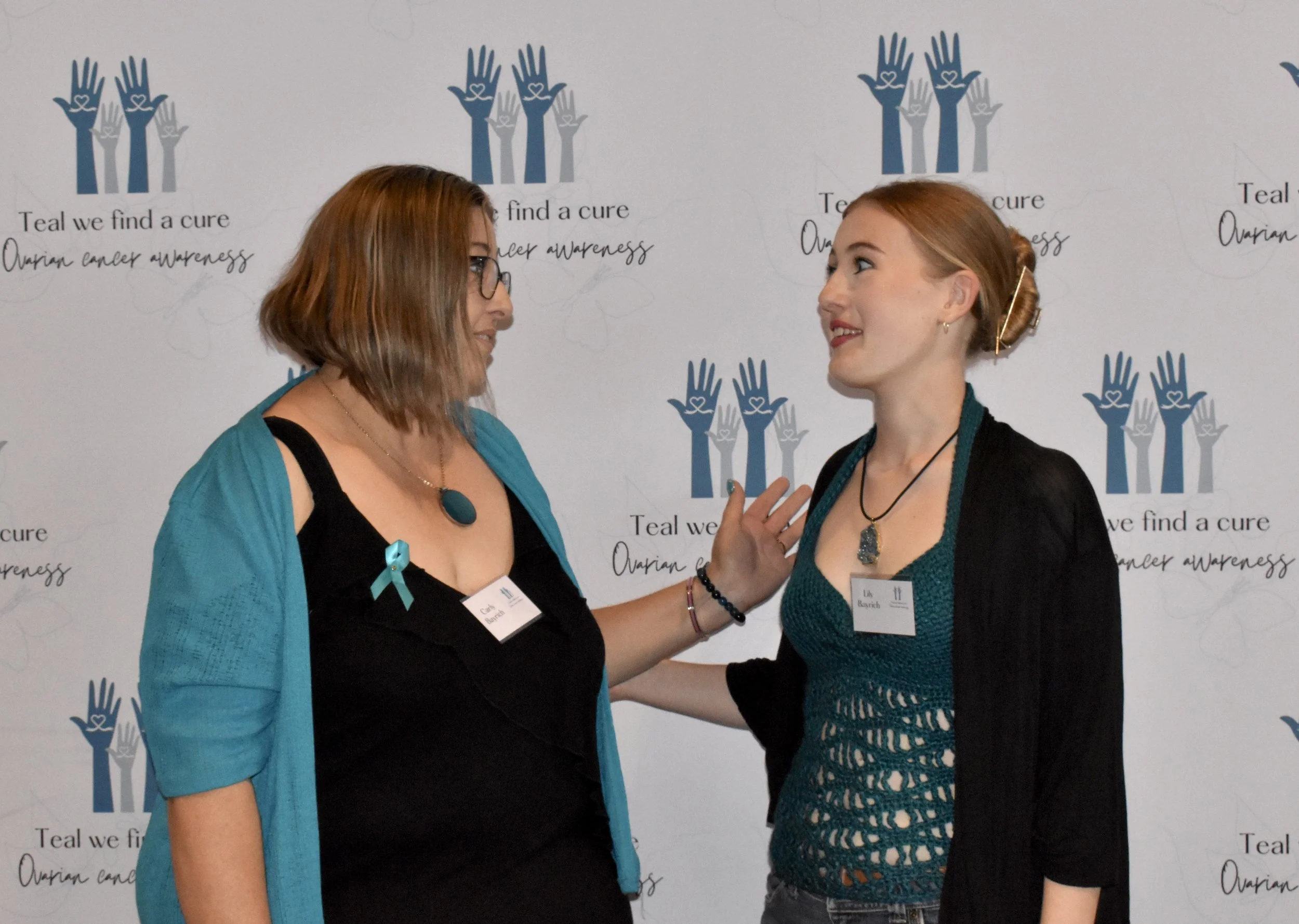 Two women having a conversation at an ovarian cancer awareness event with a backdrop that says 'Teal we find a cure, ovarian cancer awareness' and features teal-colored hands holding hearts.