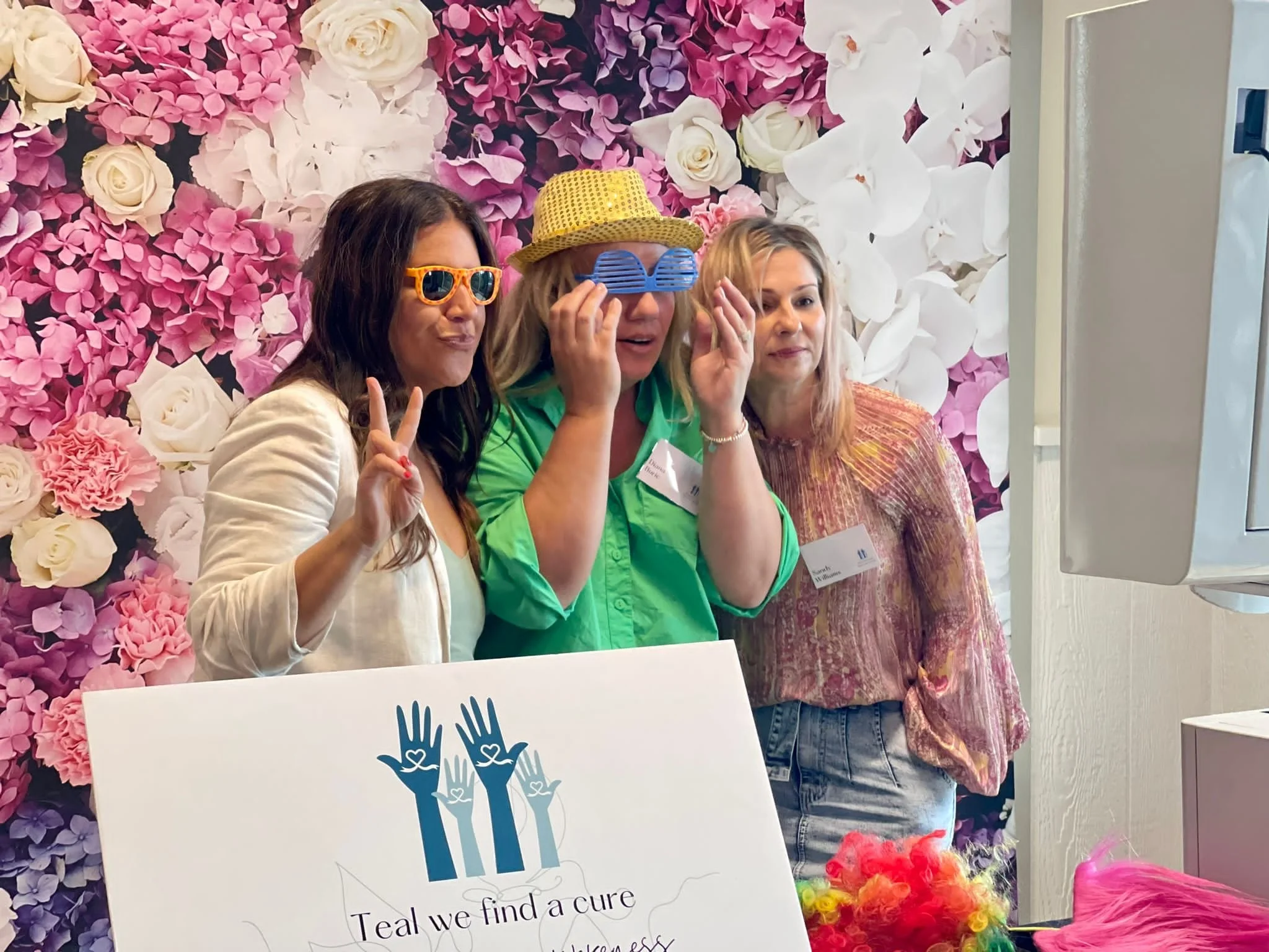 Three women posing for a photo against a pink and white floral backdrop at a breast cancer awareness event. They are wearing playful sunglasses and one woman is making a peace sign. There is a sign in front of them that reads "Teal we find a cure."
