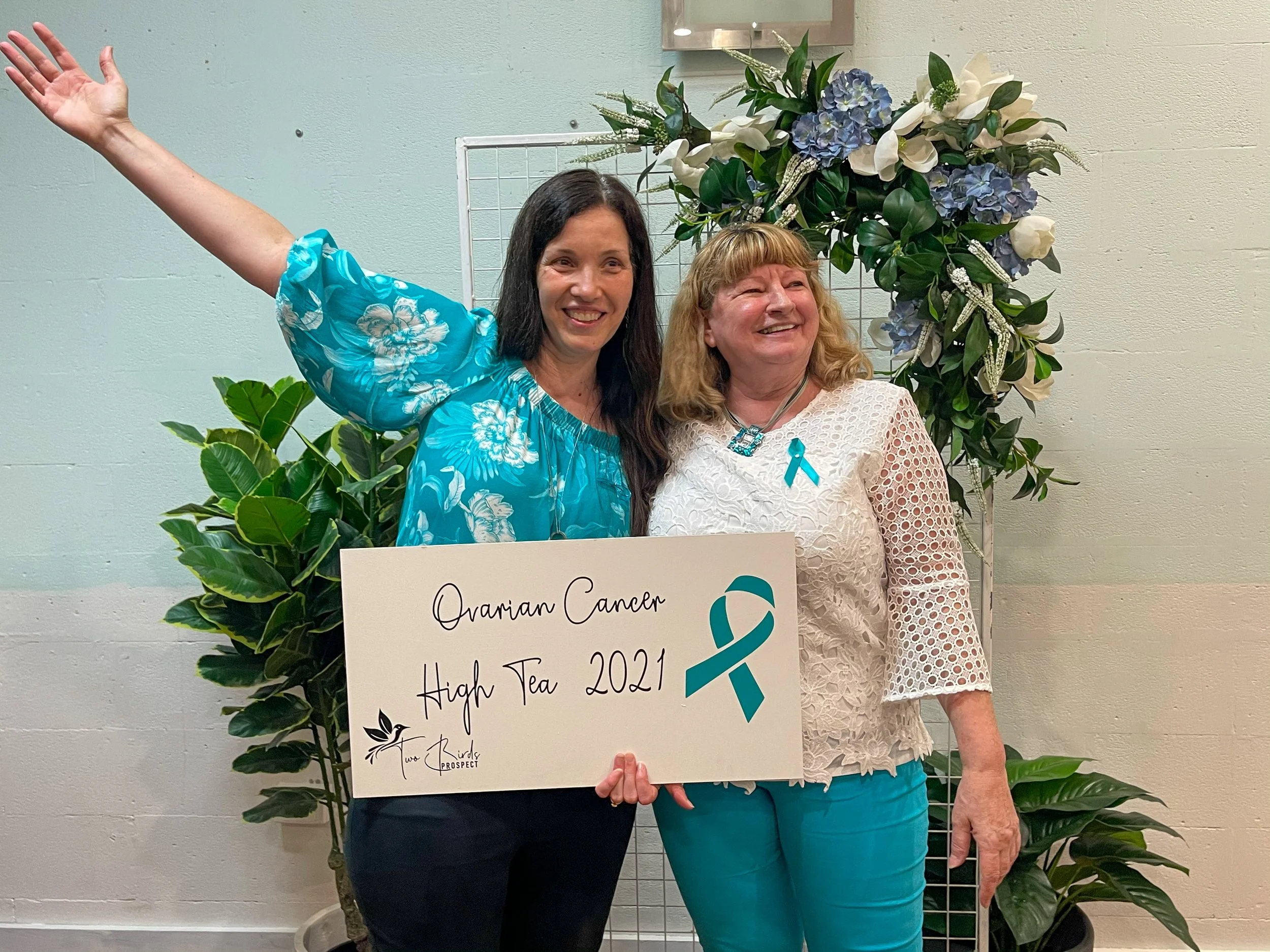 Two women smiling, one holding a large sign that says, 'Ovarian Cancer High Tea 2021' with a teal ribbon, standing in front of green plants and a floral arrangement.