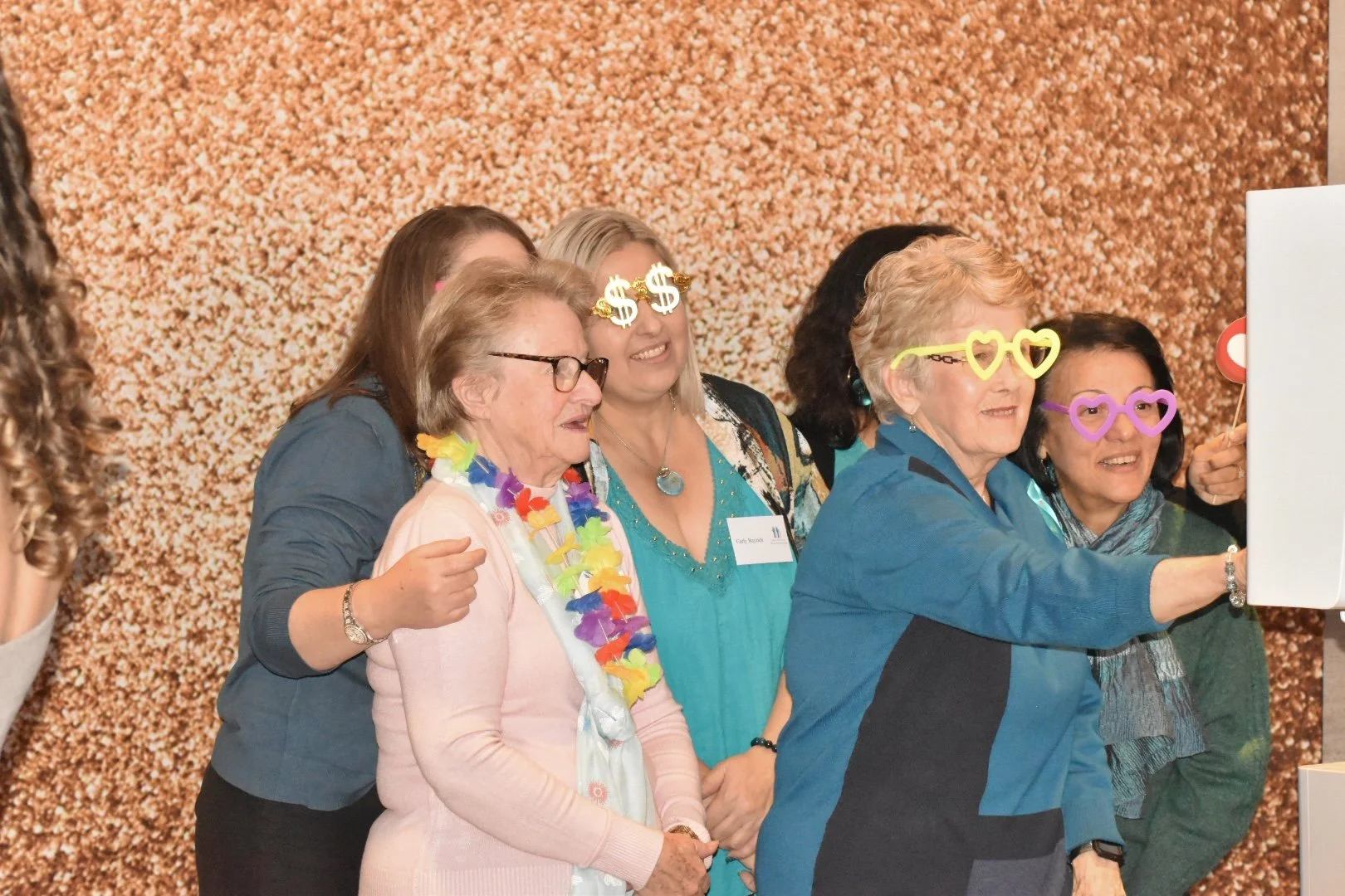 Group of women celebrating, wearing fun glasses with dollar signs, hearts, and other shapes, in front of a glittery, gold backdrop.