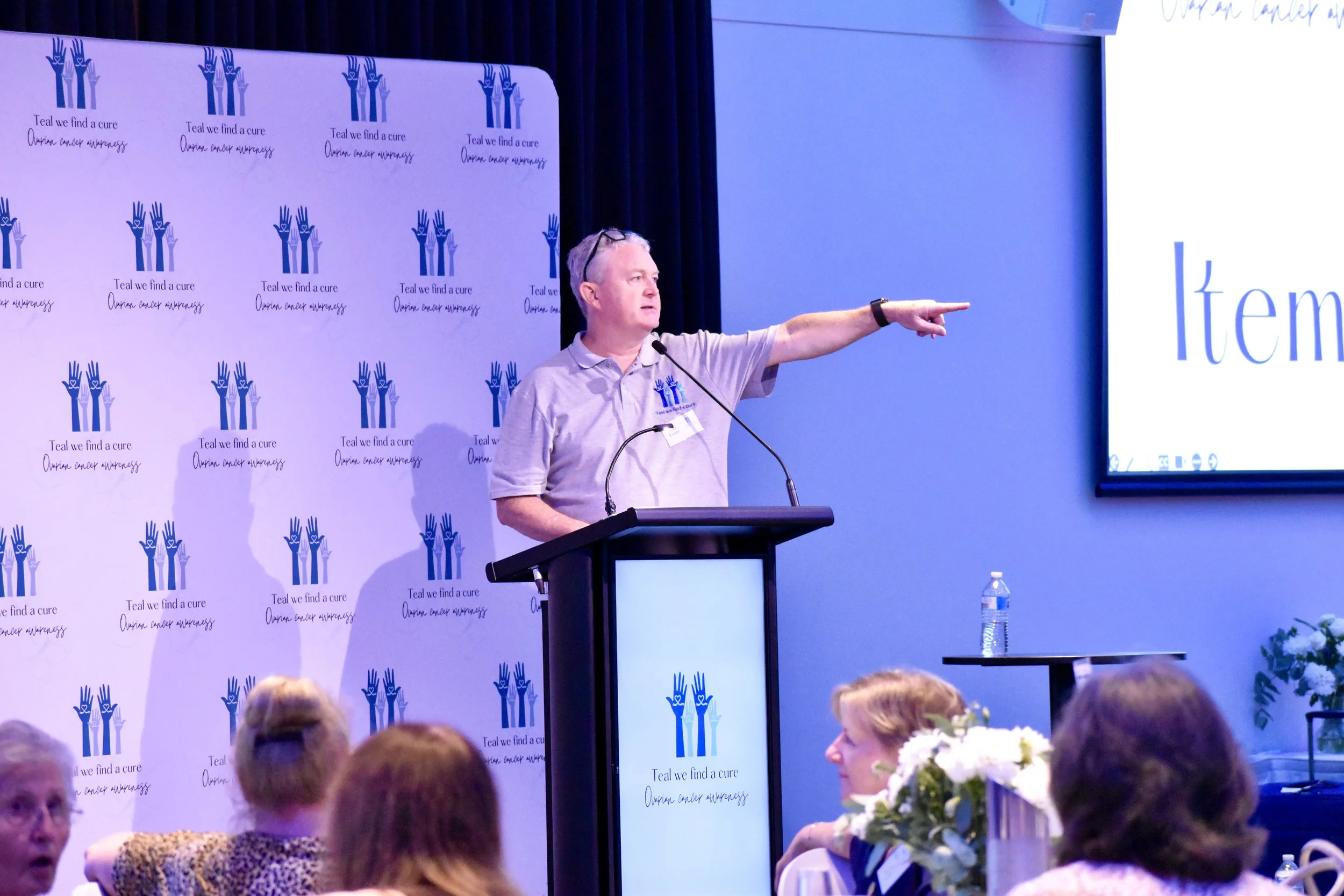 A man standing at a podium giving a presentation at a conference with a backdrop featuring a logo of three raised hands and the text 'Teal we find a cure, ovarian cancer awareness.' He is pointing towards a display screen that reads 'Item' and has a 
