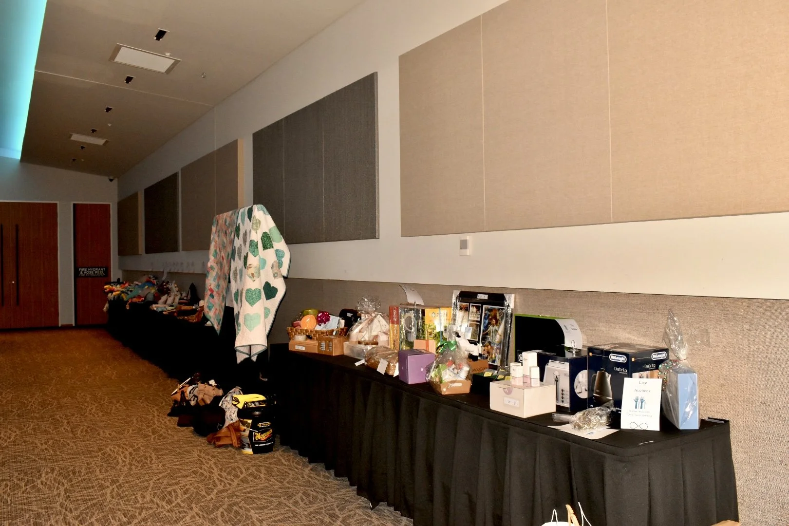 Table filled with various raffle prizes and items at an indoor event, with beige and gray wall panels in the background.