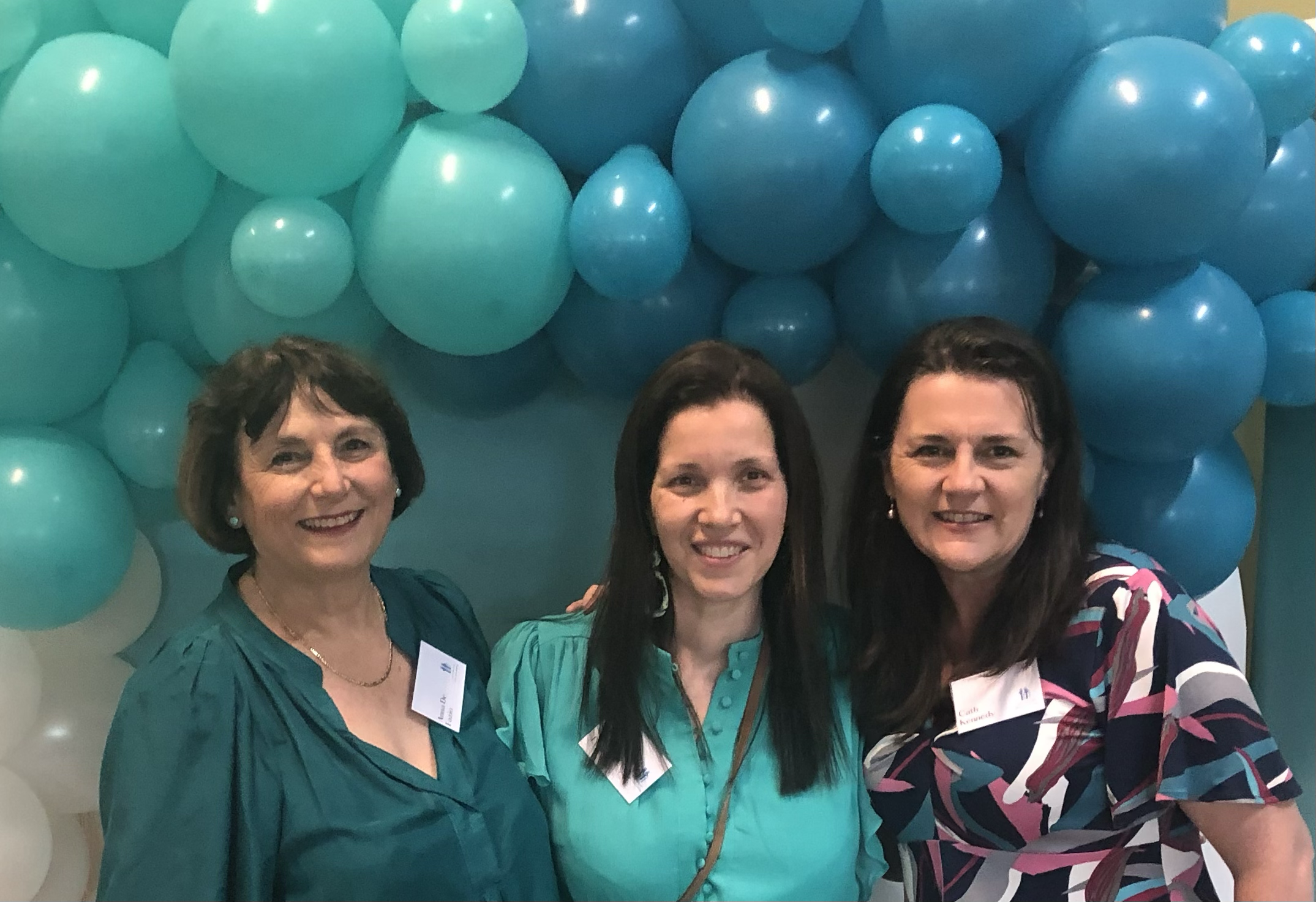 Three women smiling in front of an arrangement of teal and blue balloons at an event.