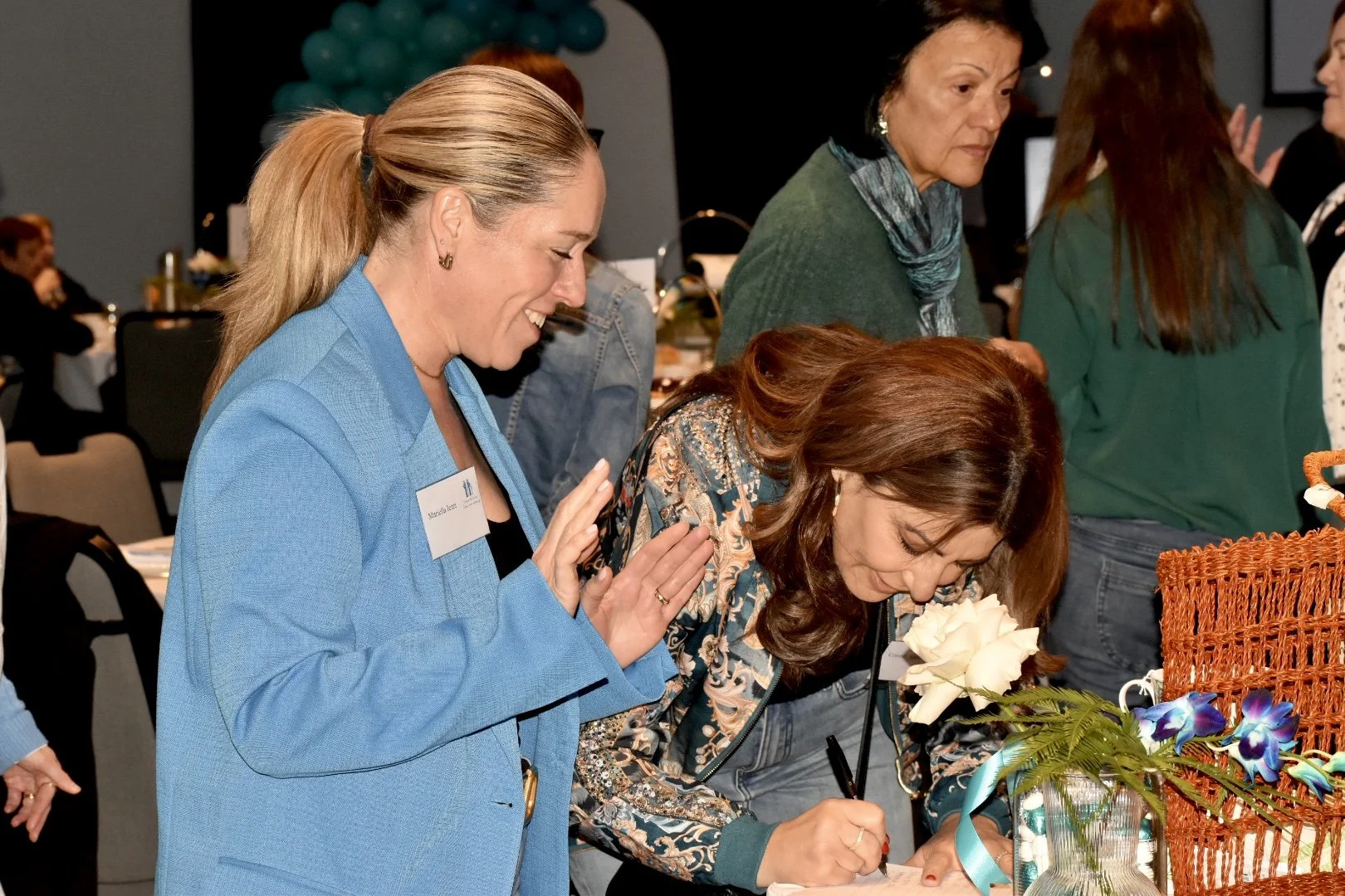 A woman with brown hair is signing a book or paper, while another woman with blonde hair, wearing a blue jacket, is watching and smiling. There is a floral arrangement in front of the signing woman and several people and tables in the background.