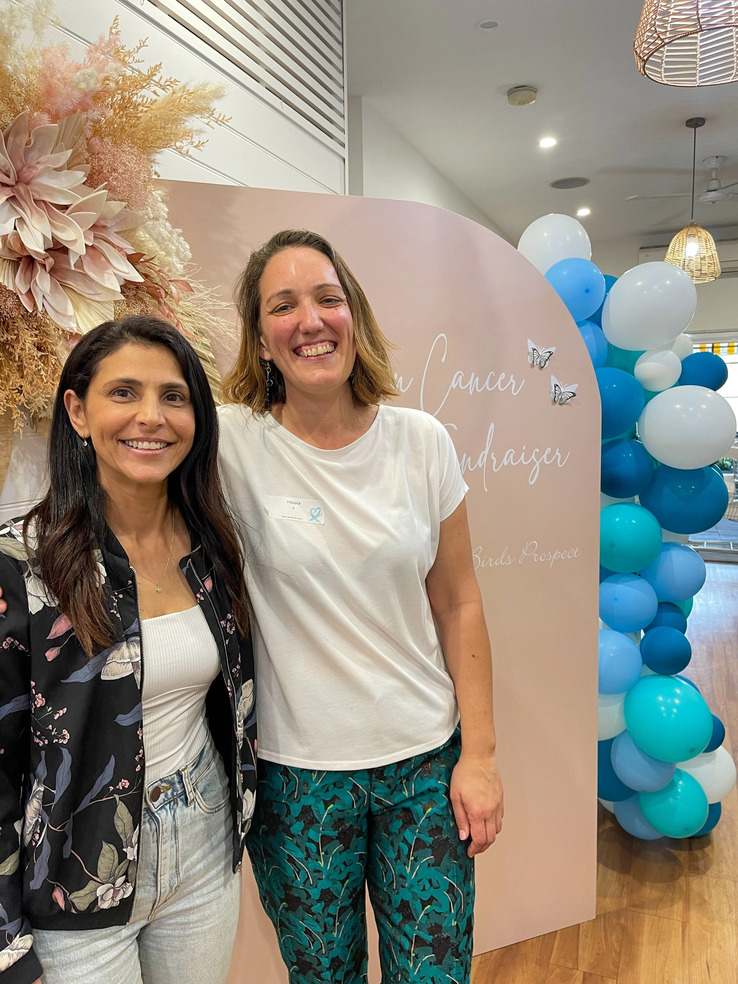Two women smiling at a breast cancer fundraiser event, in front of a pink backdrop decorated with flowers and butterflies, with a balloon arch in shades of blue and white.