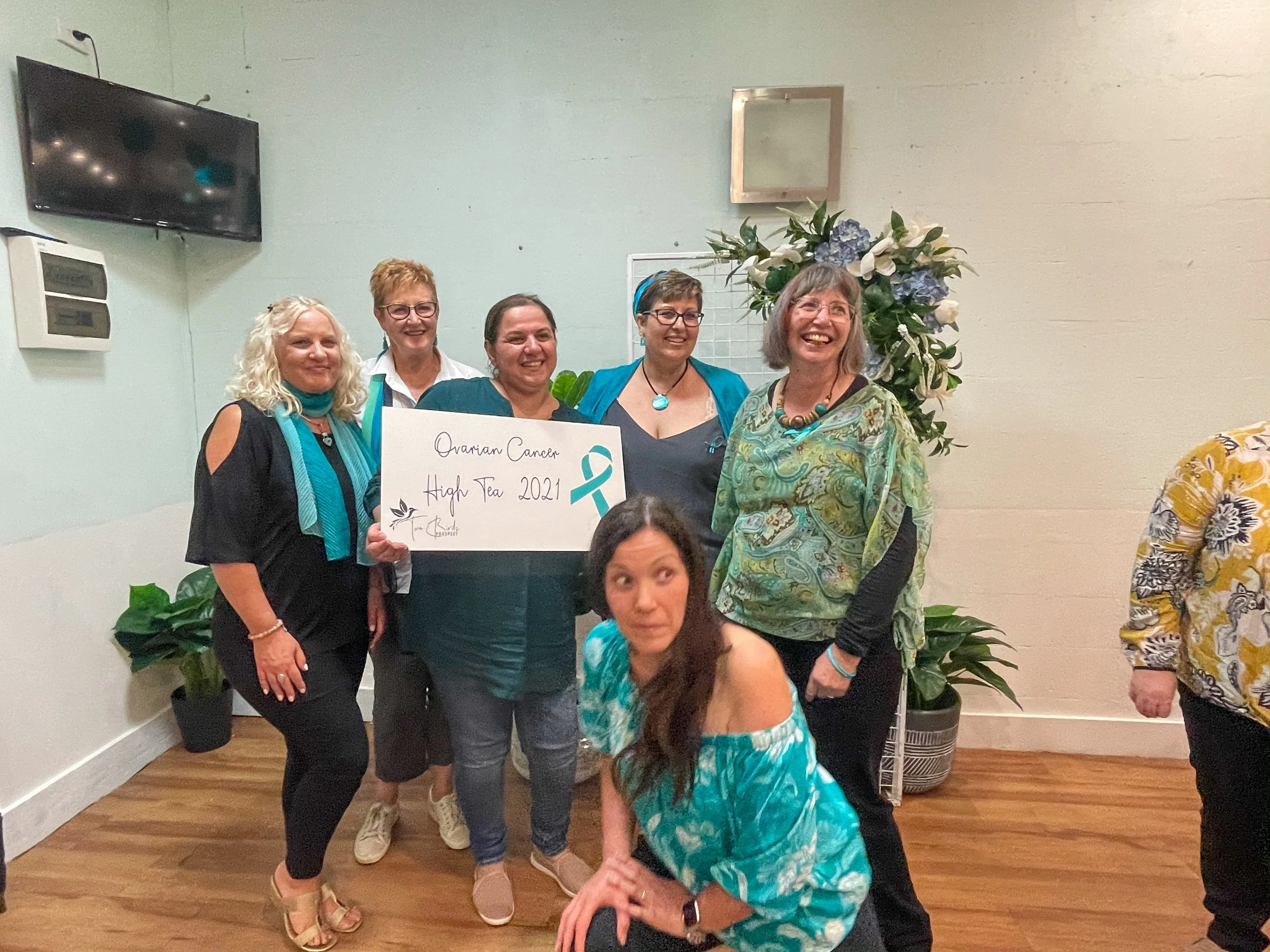 Group of women celebrating ovarian cancer awareness at High Tea 2021, holding a sign with a teal ribbon and text, standing near potted plants and floral arrangements.