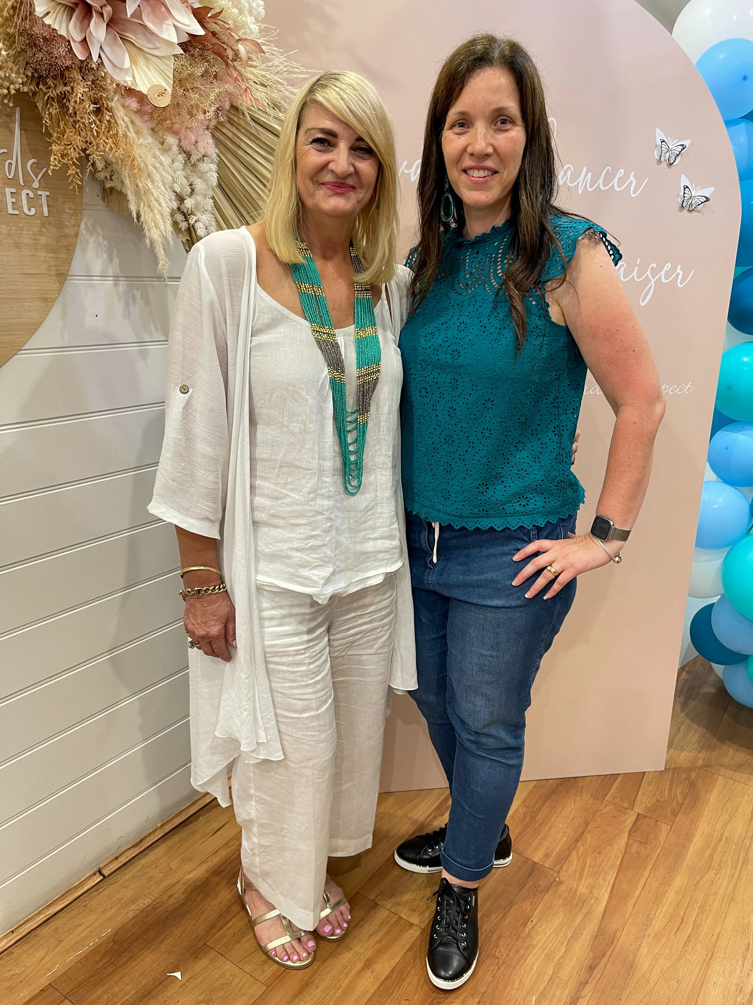 Two women standing together at a celebratory event with a pink backdrop and blue balloons. The woman on the left is wearing a white outfit with gold sandals and jewelry, and the woman on the right is wearing a teal top, jeans, and sneakers.