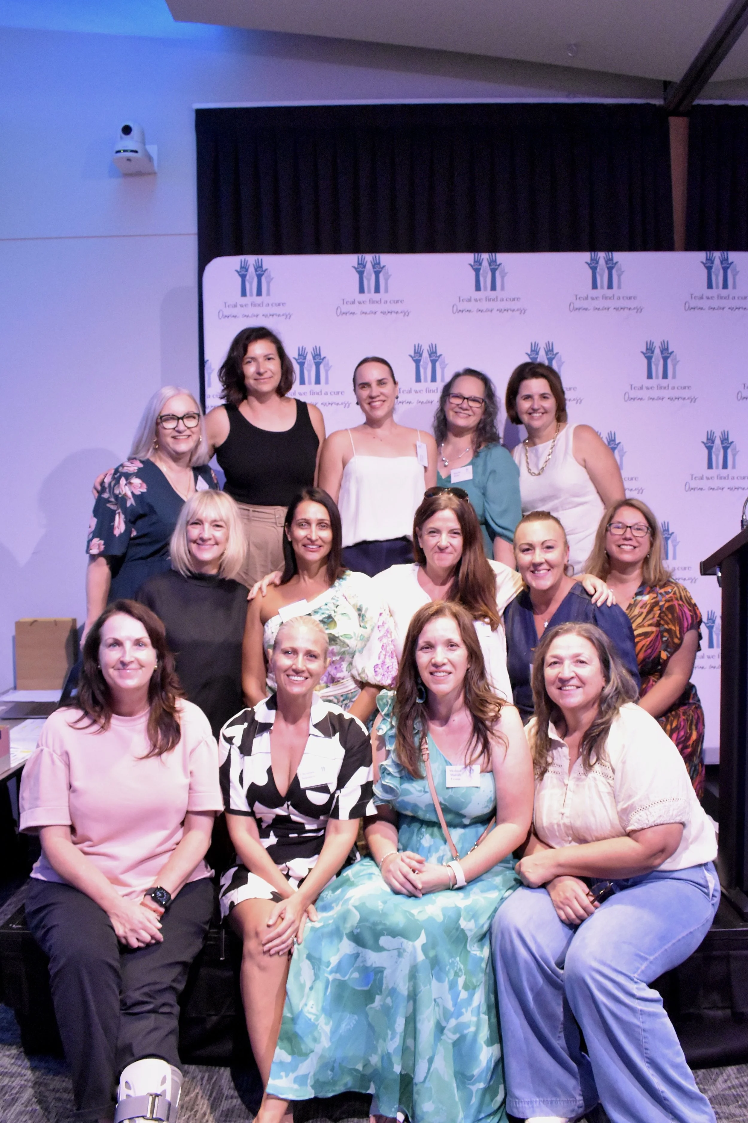 Group of women smiling for a photo at an event with a backdrop that reads 'Tell us find a cure, donate, donate, donate' with blue handprints.