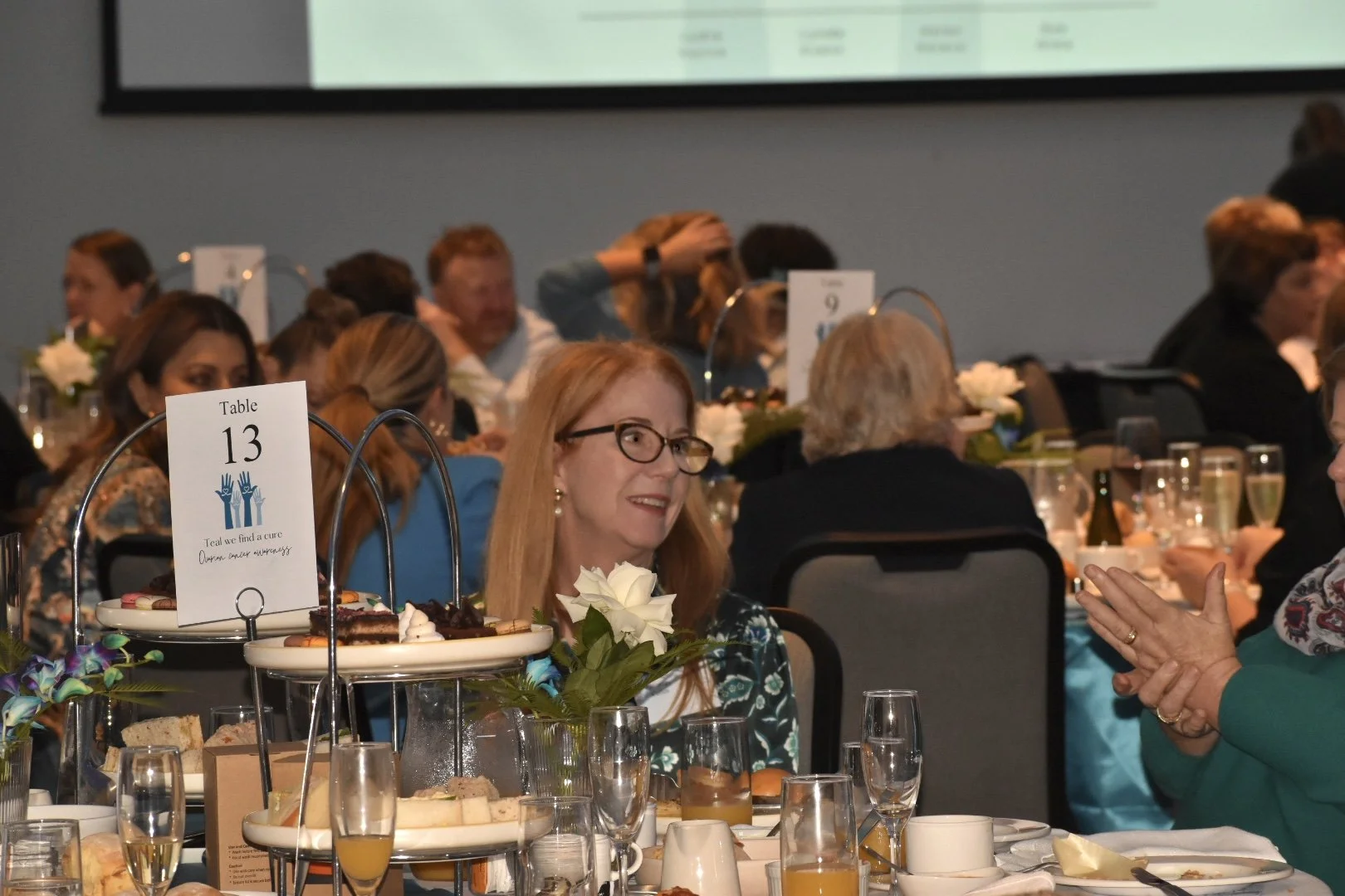 A banquet hall filled with people seated at decorated tables during a formal event. The table has a sign for Table 13, flowers, and an assortment of food and drinks. People are engaged in conversation, with some clapping and others listening attentiv
