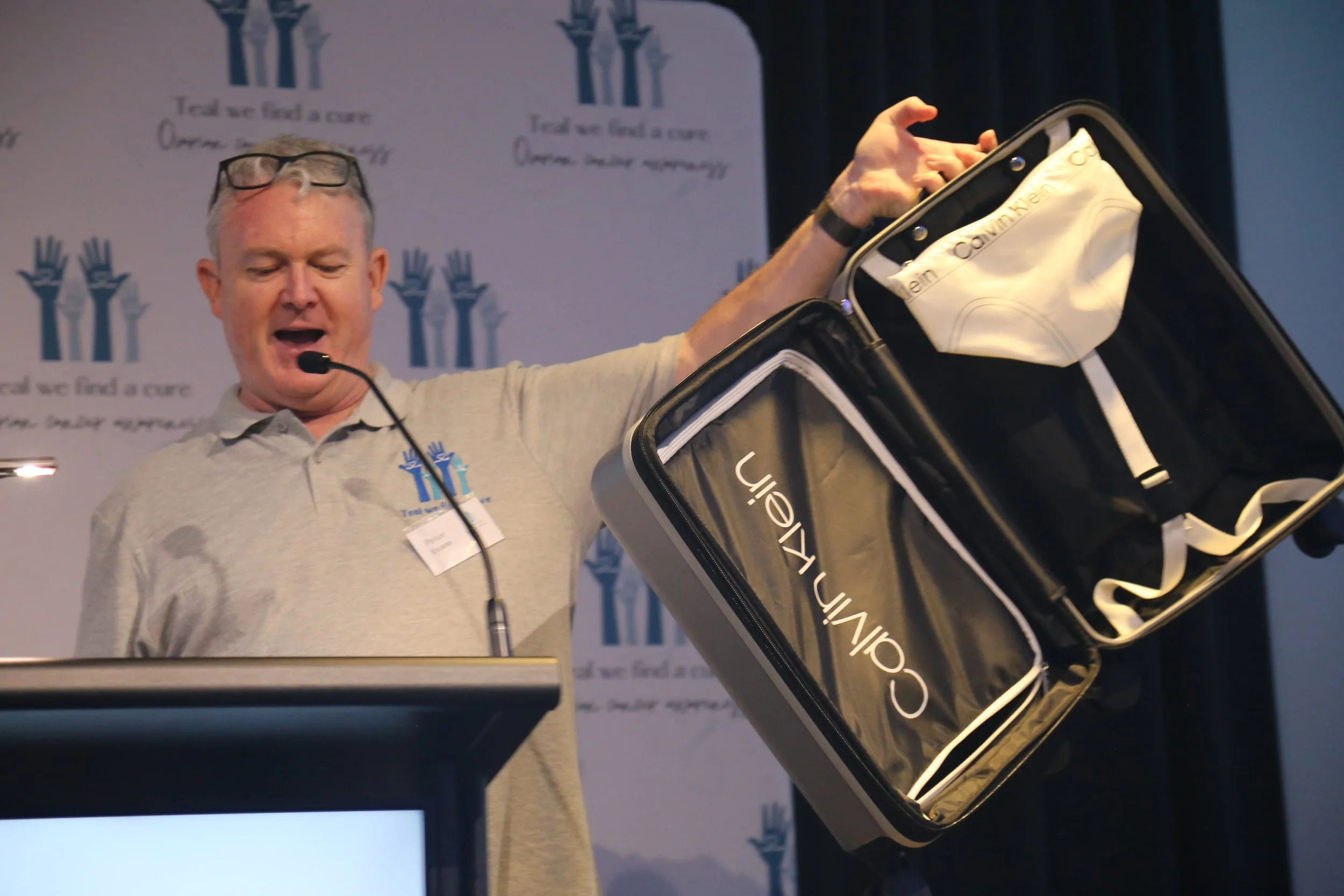A man is holding a black and white Calvin Klein suitcase with a white bag inside, at a conference or event with a backdrop featuring the text 'Trust we find a cure' and the logo of a charity organization.