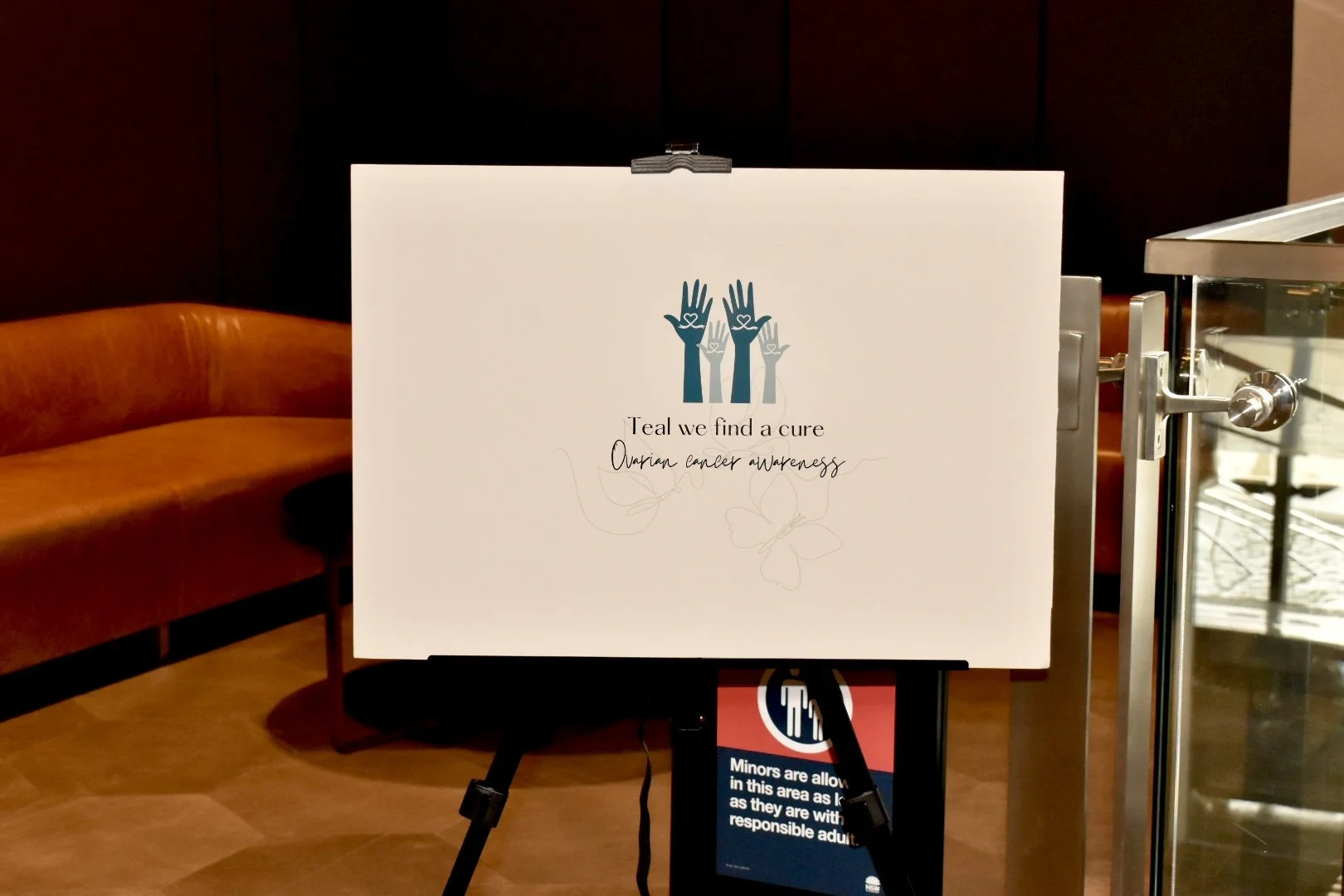 Sign on an easel promoting ovarian cancer awareness with stylized illustrations of hands and butterflies, and a message about finding a cure.