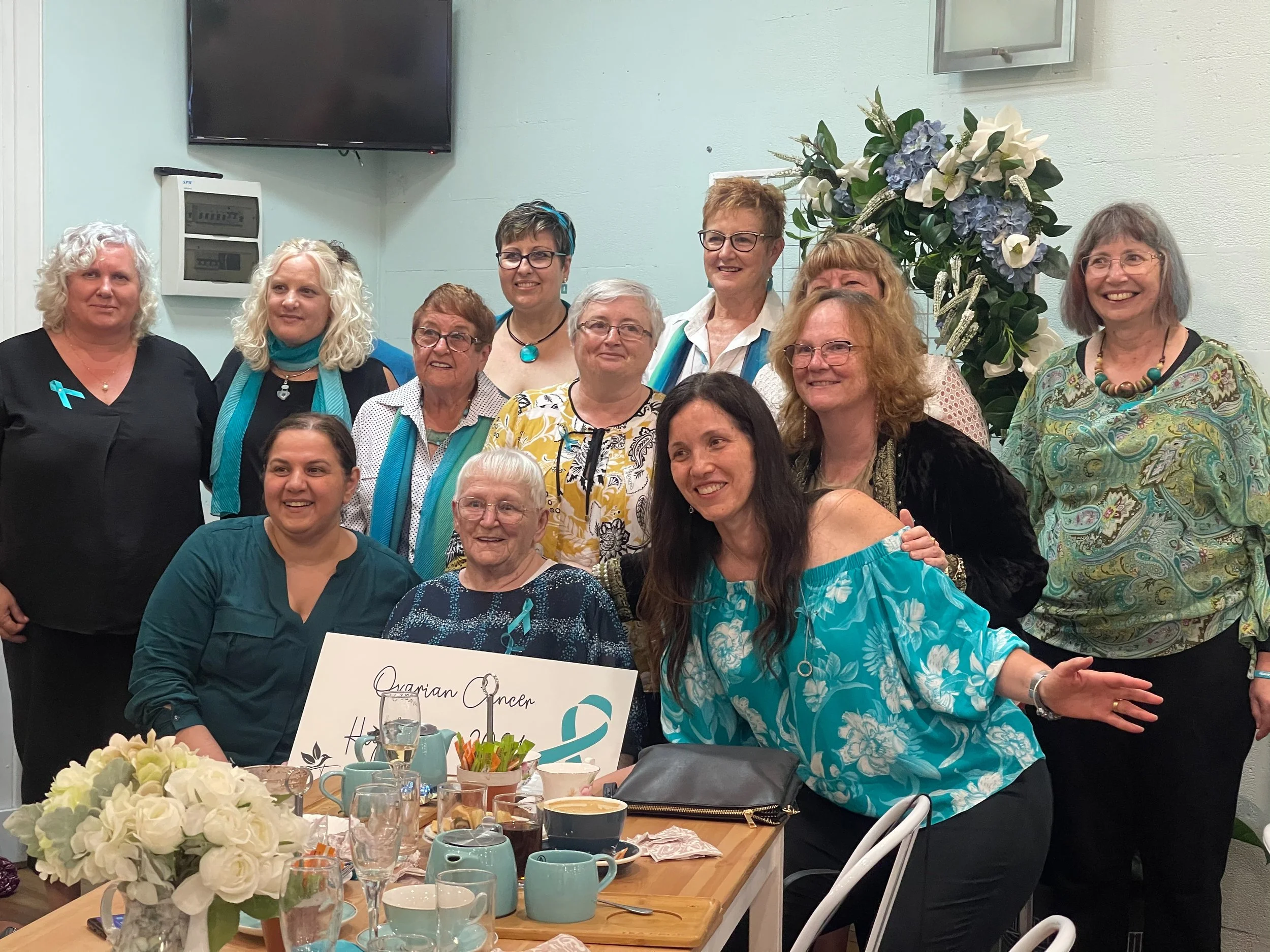 A group of women gathered around a table with flowers and food, smiling for a photo in a celebration setting. There is a flower arrangement and a whiteboard with writing on it in front of the elderly woman seated in the center.