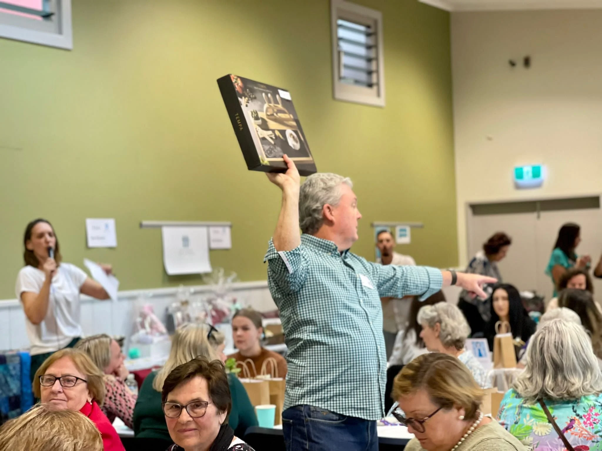 A man wearing a checkered shirt is standing with his arm raised, holding a box. He is in a room filled with seated women, some with glasses, and a woman with a microphone is standing in the background.