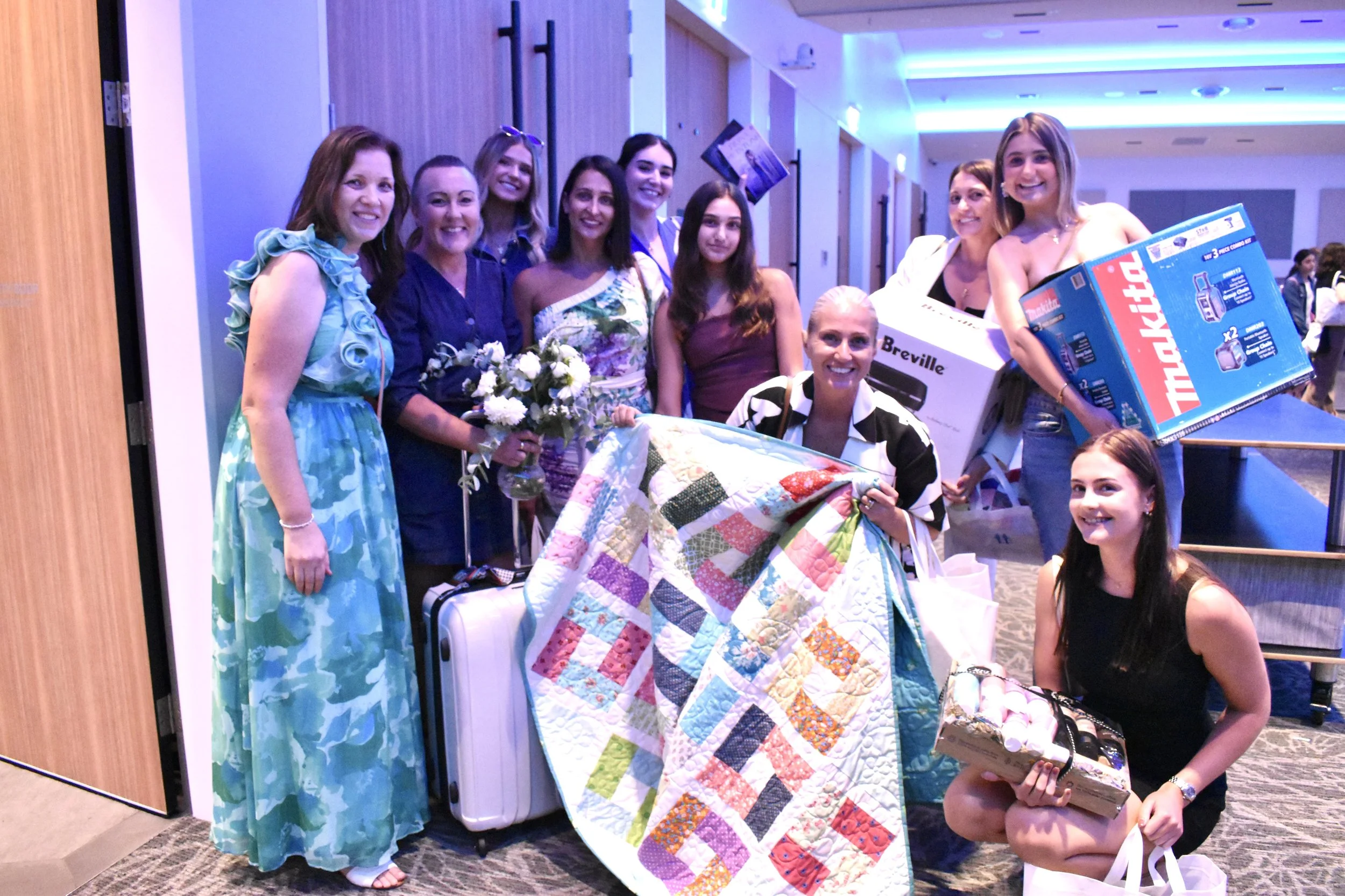 A group of women and girls posing together indoors, some holding gift boxes, flowers, and a colorful quilt, with bright lighting and a modern interior background.
