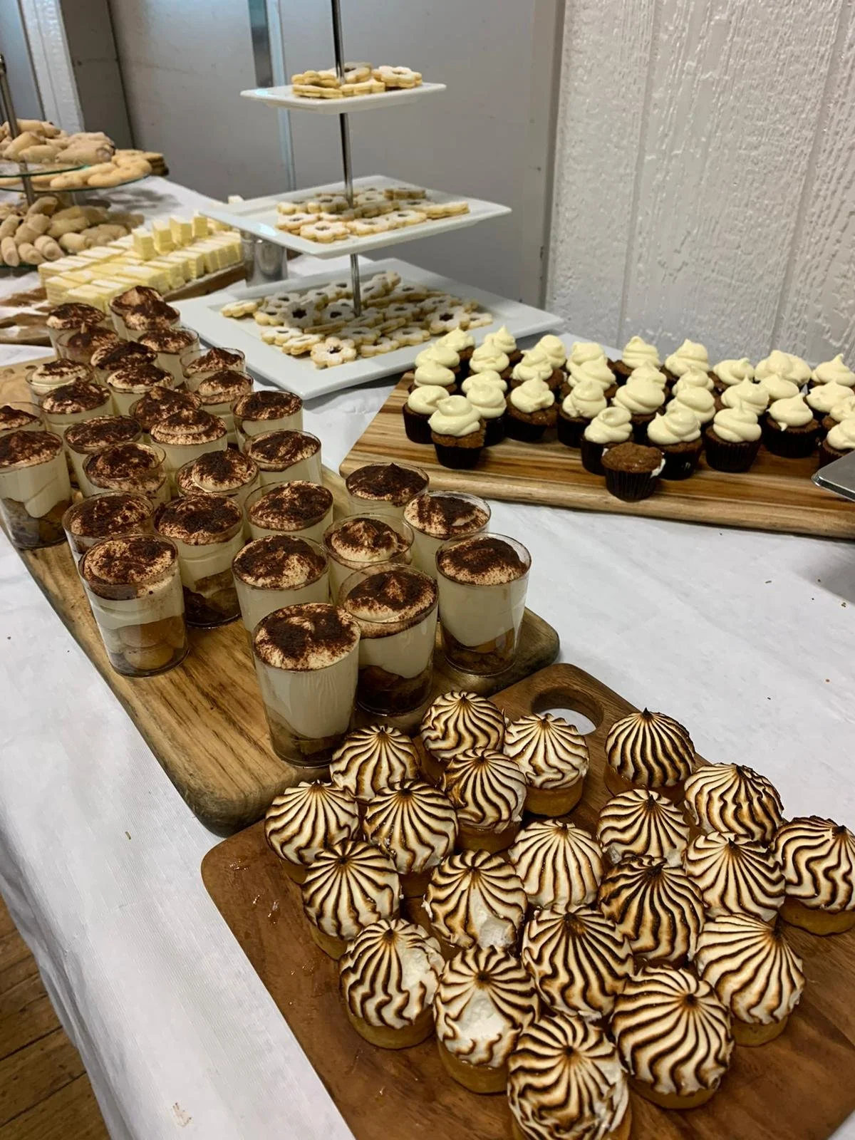 Assorted desserts on a buffet table, including tiramisu in glasses, decorated cookies, cupcakes with white frosting, and sprinkled cookies.