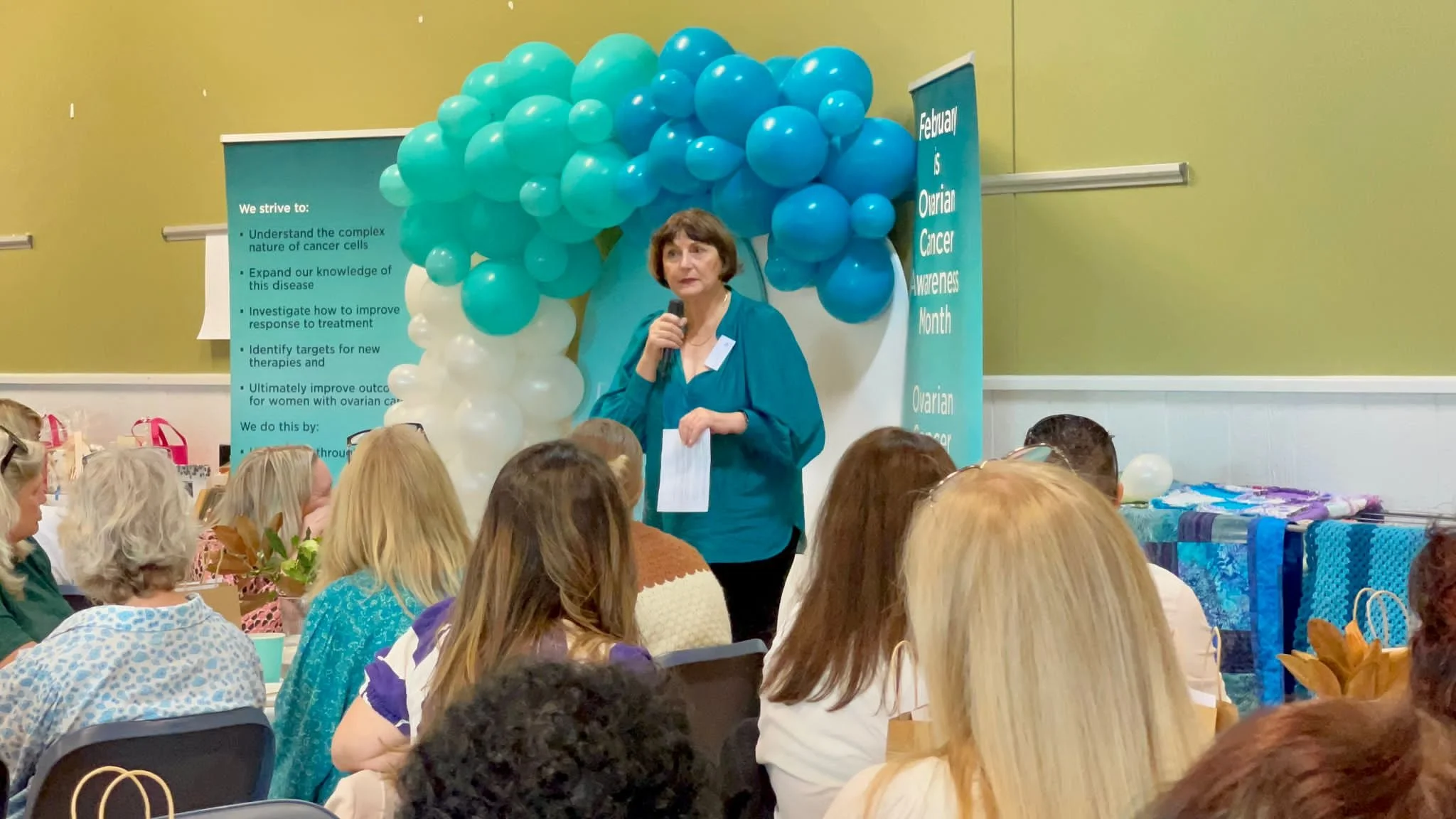 A woman speaking into a microphone at a breast cancer awareness event, with a teal and white balloon arrangement behind her and a teal banner indicating February is Ovarian Cancer Awareness Month. Attendees sit in front, listening.
