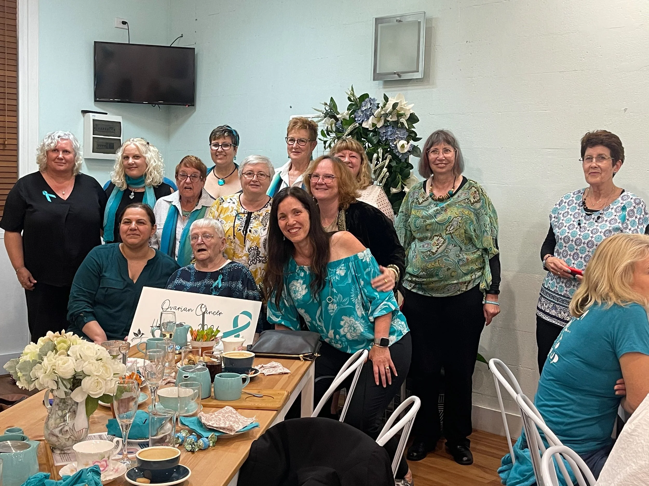 A group of women gathered around a dining table at a women’s health event, with teal ribbons and flowers, some wearing teal accessories, and a sign that reads 'Ovarian Cancer'.