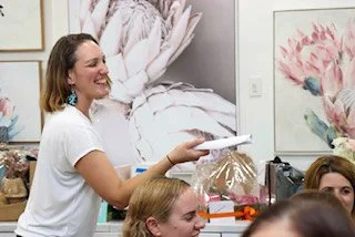 A woman handing out paper or flyers to a group of people in an indoor setting, with artwork and a bed visible in the background.