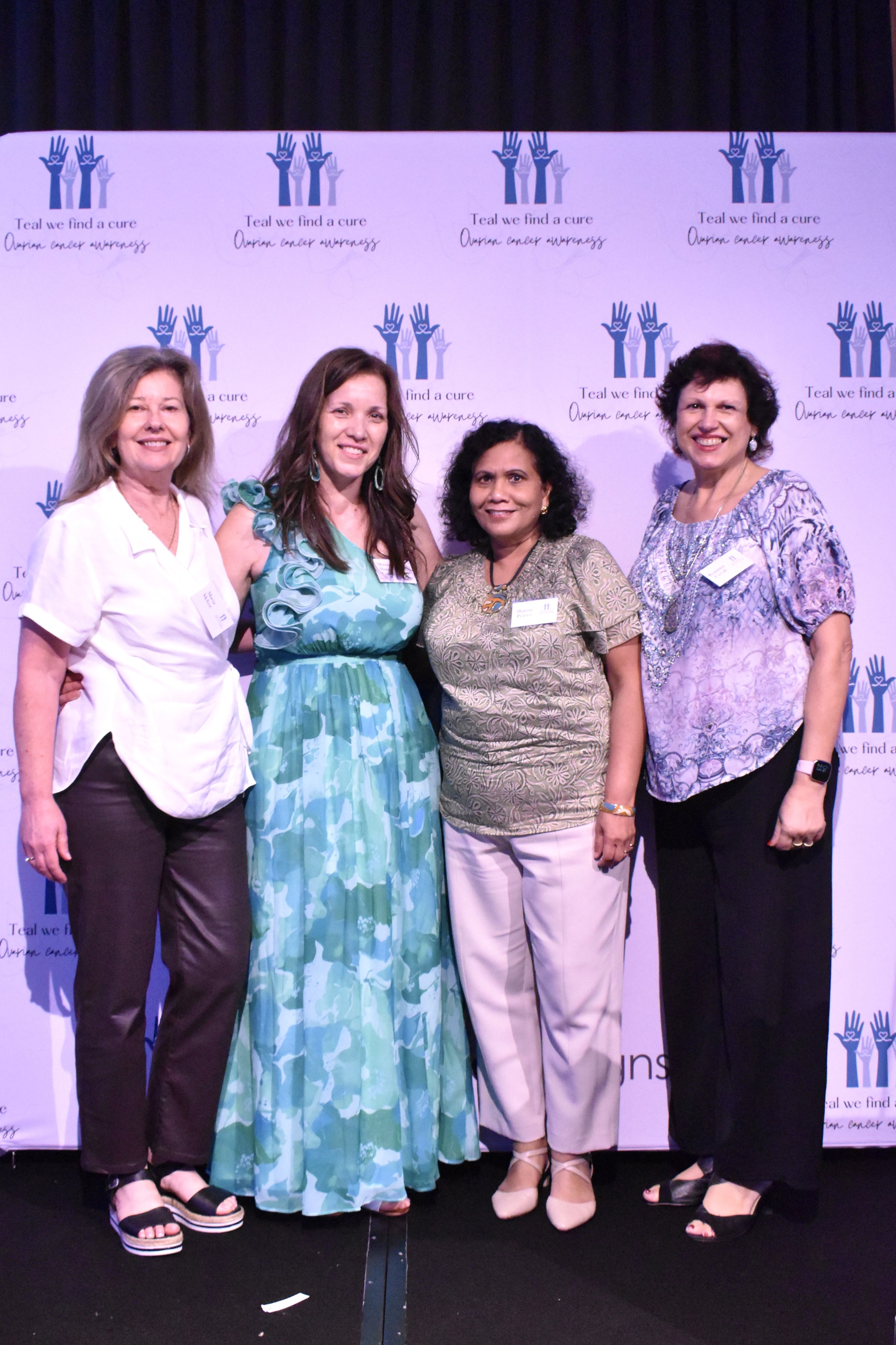 Four women standing together at an event with a backdrop that reads 'Teal we find a cure, ovarian cancer awareness' with a blue and purple hand logo.