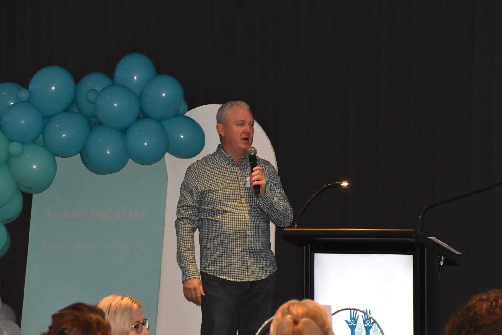 A man with gray hair, wearing a checkered shirt, is speaking into a microphone on stage during a presentation. Behind him, there are blue balloons and a podium with a screen displaying a logo, with some attendees visible in the foreground.