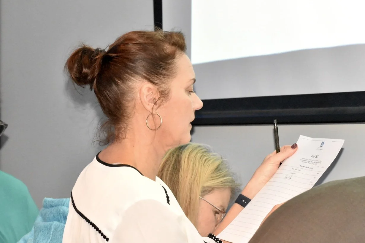 A woman with reddish-brown hair tied in a bun holding a notepad and writing, seated in a meeting or classroom setting.