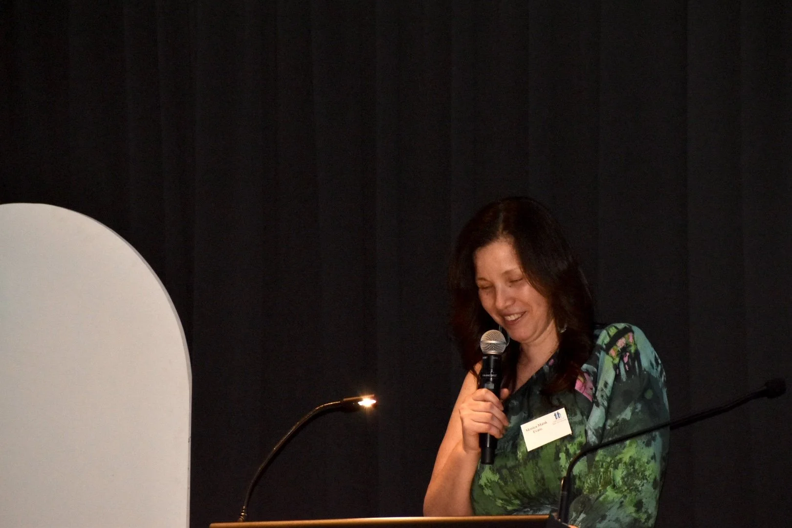 A woman with dark hair wearing a colorful dress, smiling and speaking into a microphone.