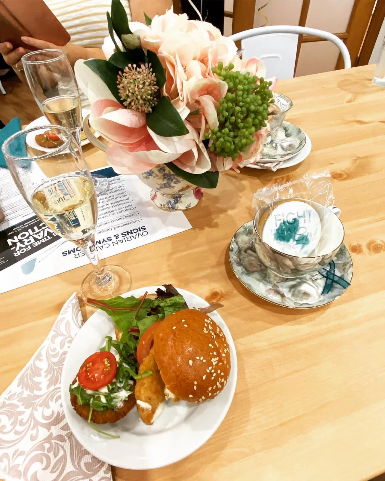 A table setting with a floral centerpiece, two glasses of white wine, a cup and saucer with a tea bag, and a plate with a sandwich and a salad.