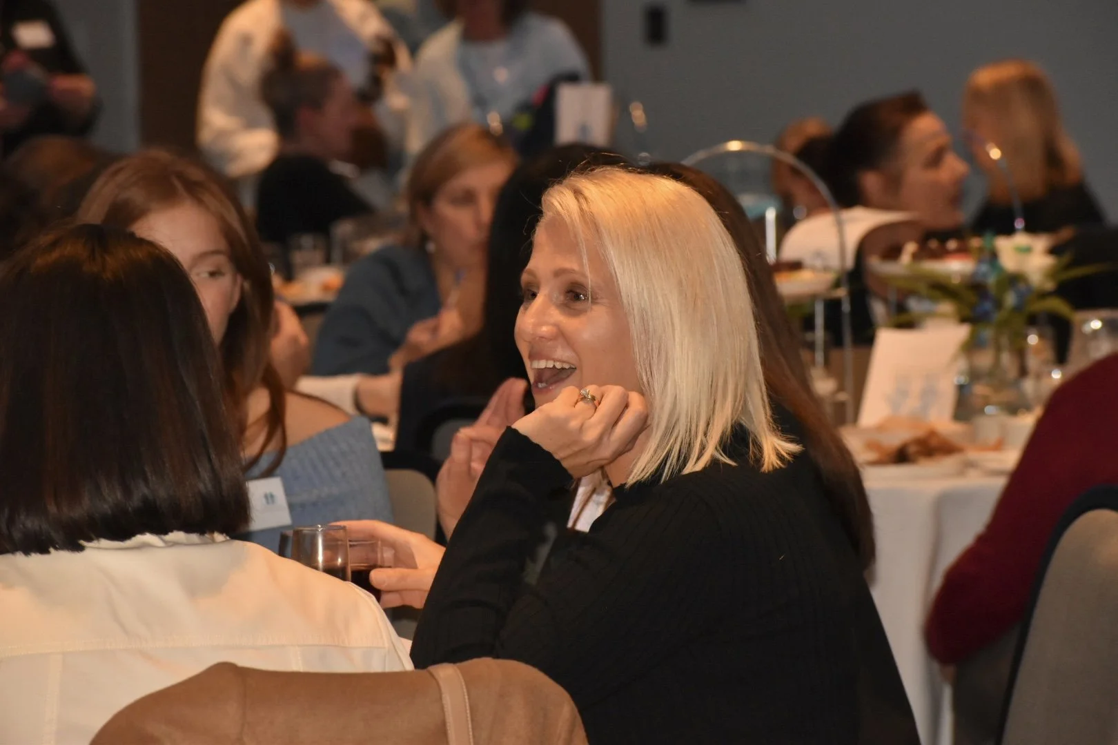 A woman with shoulder-length blond hair, smiling and talking to someone at a formal event with several other women seated at tables in the background.