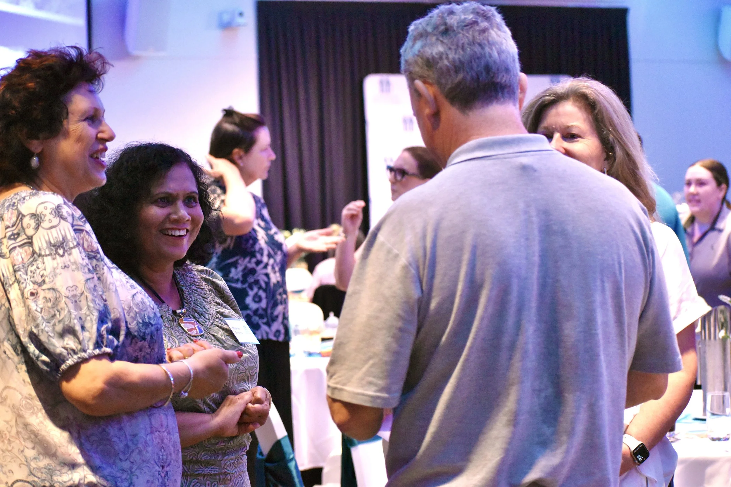 Group of people engaging in conversation at a social event or conference, with some smiling and others listening attentively, in a well-lit room with tables and chairs.