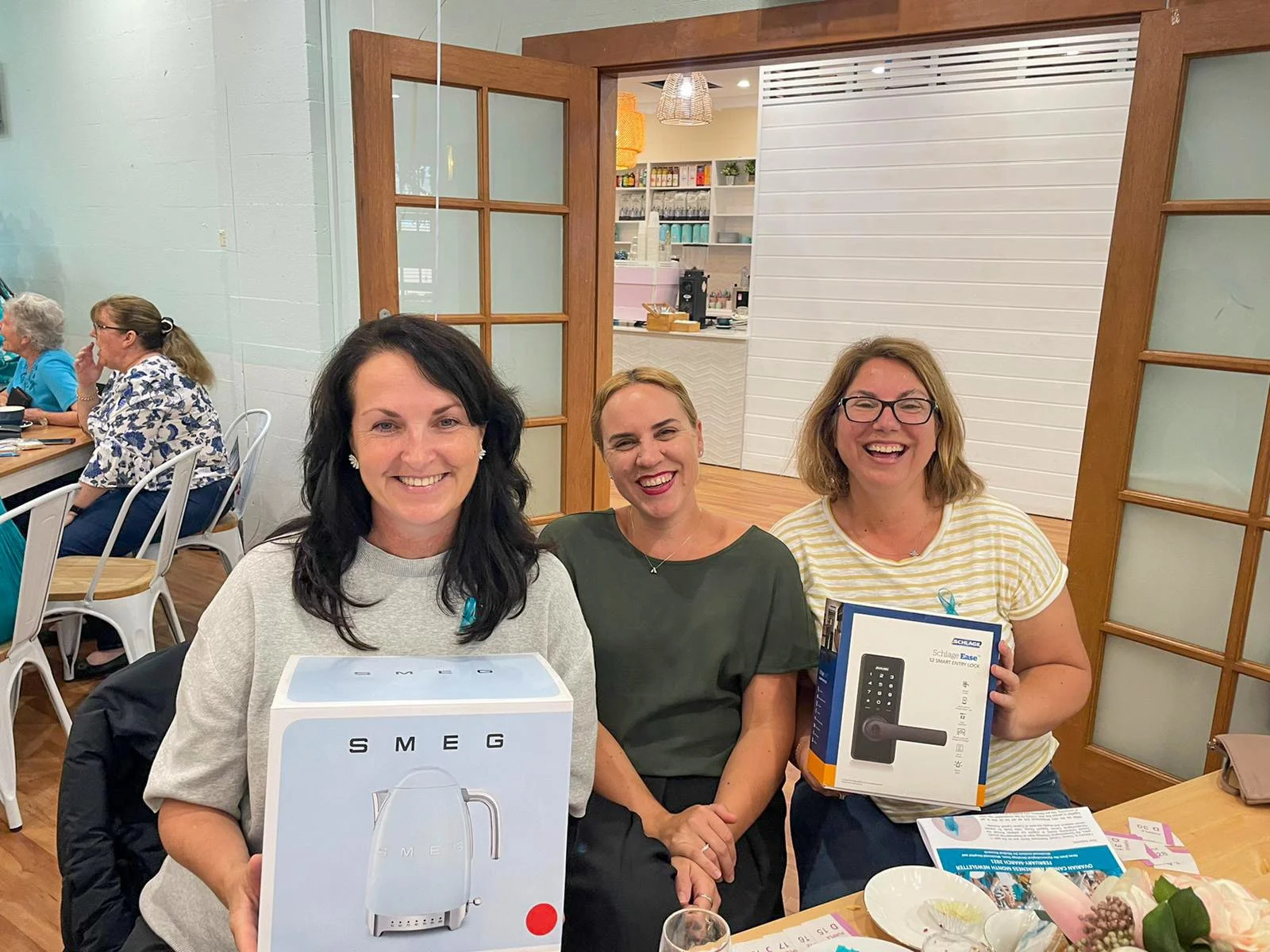 Three women in a restaurant or cafe, smiling at the camera, holding boxes of small appliances, with other women in the background, and a table with paper and flowers in the foreground.