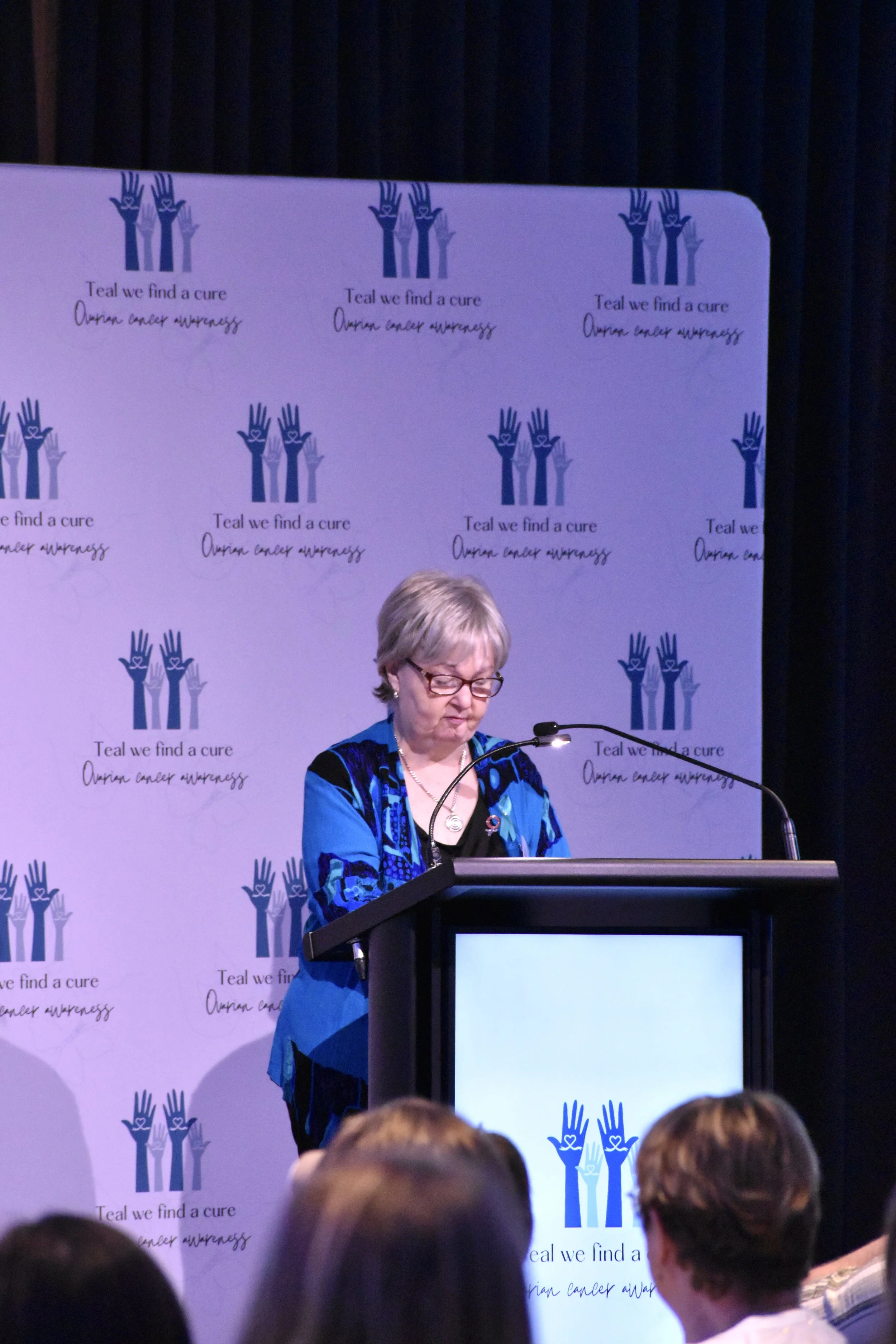 A woman standing at a podium speaking to an audience, with a backdrop that has the logo and slogan for ovarian cancer awareness.