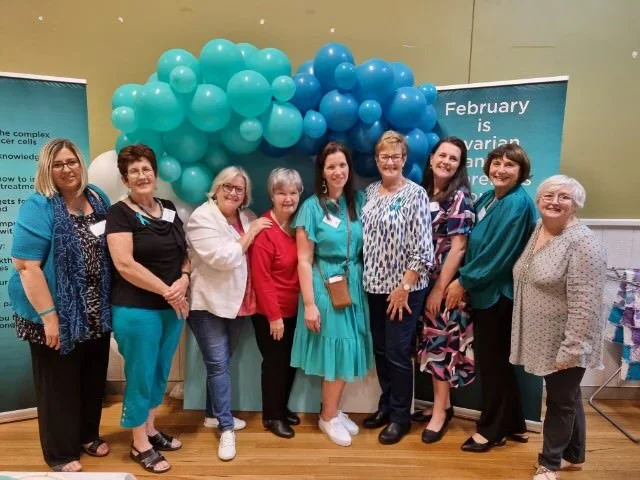 A group of nine women standing together in front of a cluster of teal and blue balloons at an indoor event.