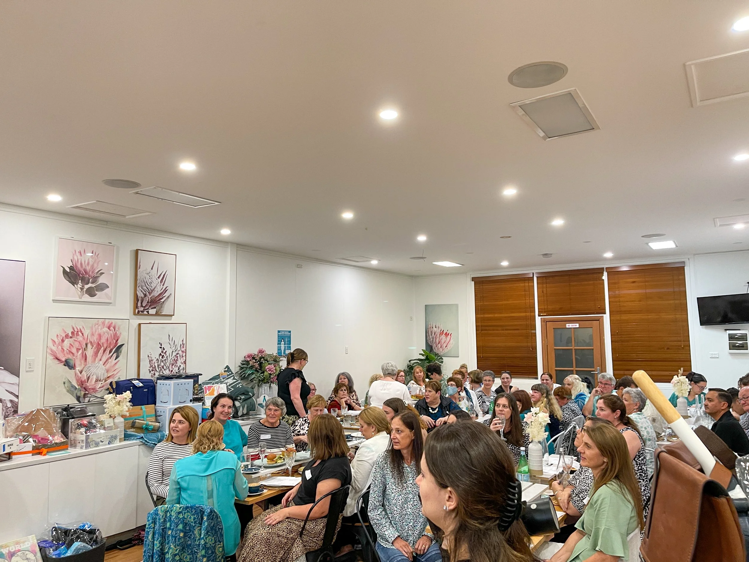 Large group of women gathered in a well-lit room for a social event, sitting at tables with food and drinks, with floral artwork on the walls and wooden blinds on the windows.