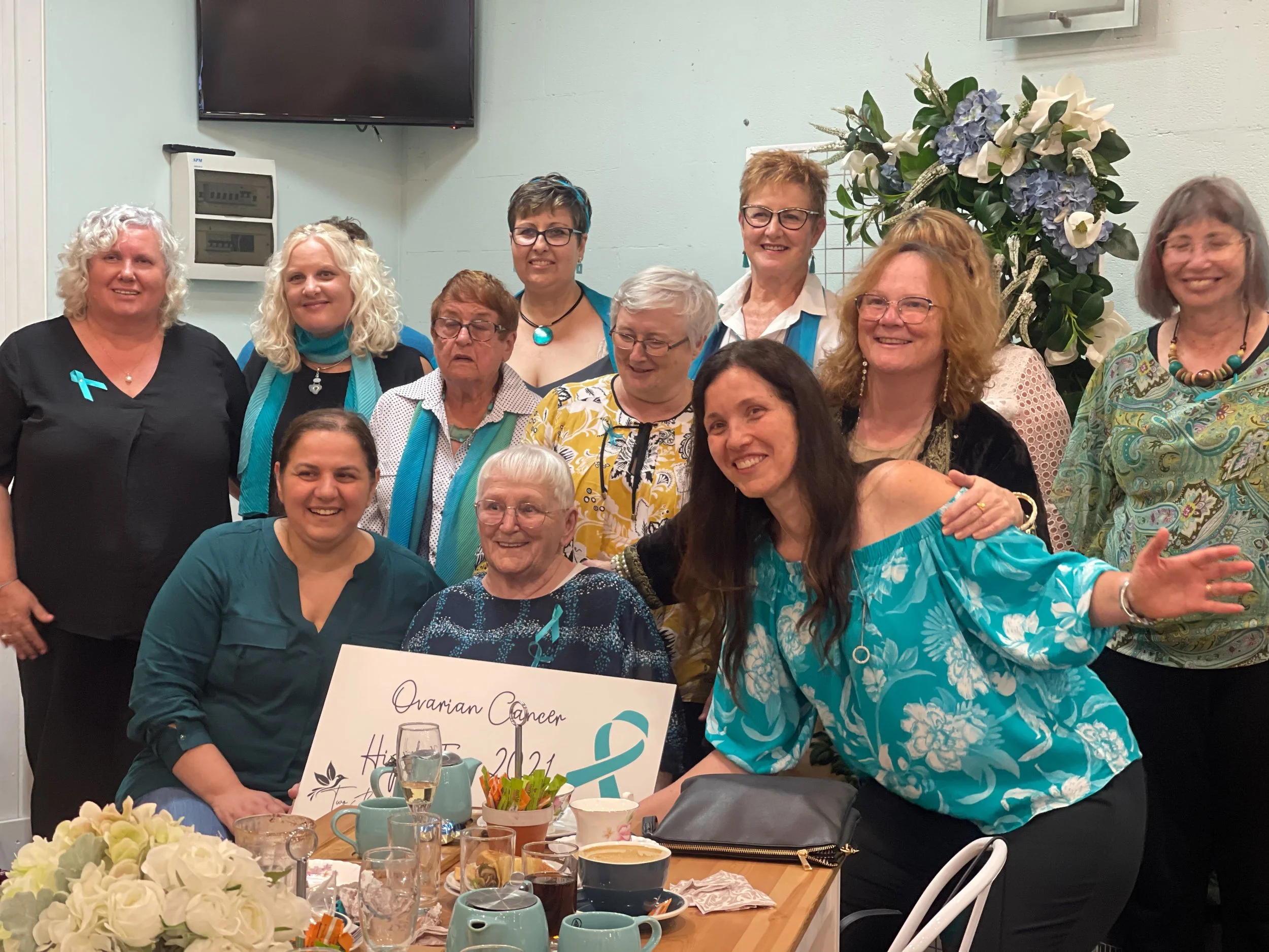 Group of women gathered for a celebration, some holding a sign that reads 'Ovarian Cancer Hope 2021,' with floral decorations and a table set with drinks and cups.