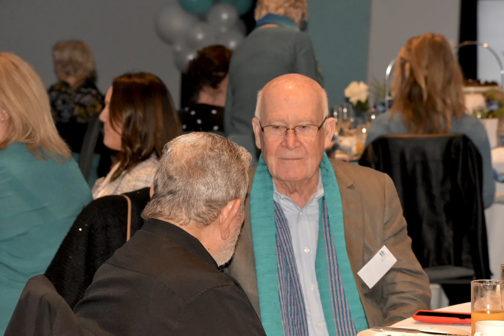 An elderly man with glasses and a blue scarf talking to a man with gray hair at a formal gathering, with other guests seated at tables in the background.