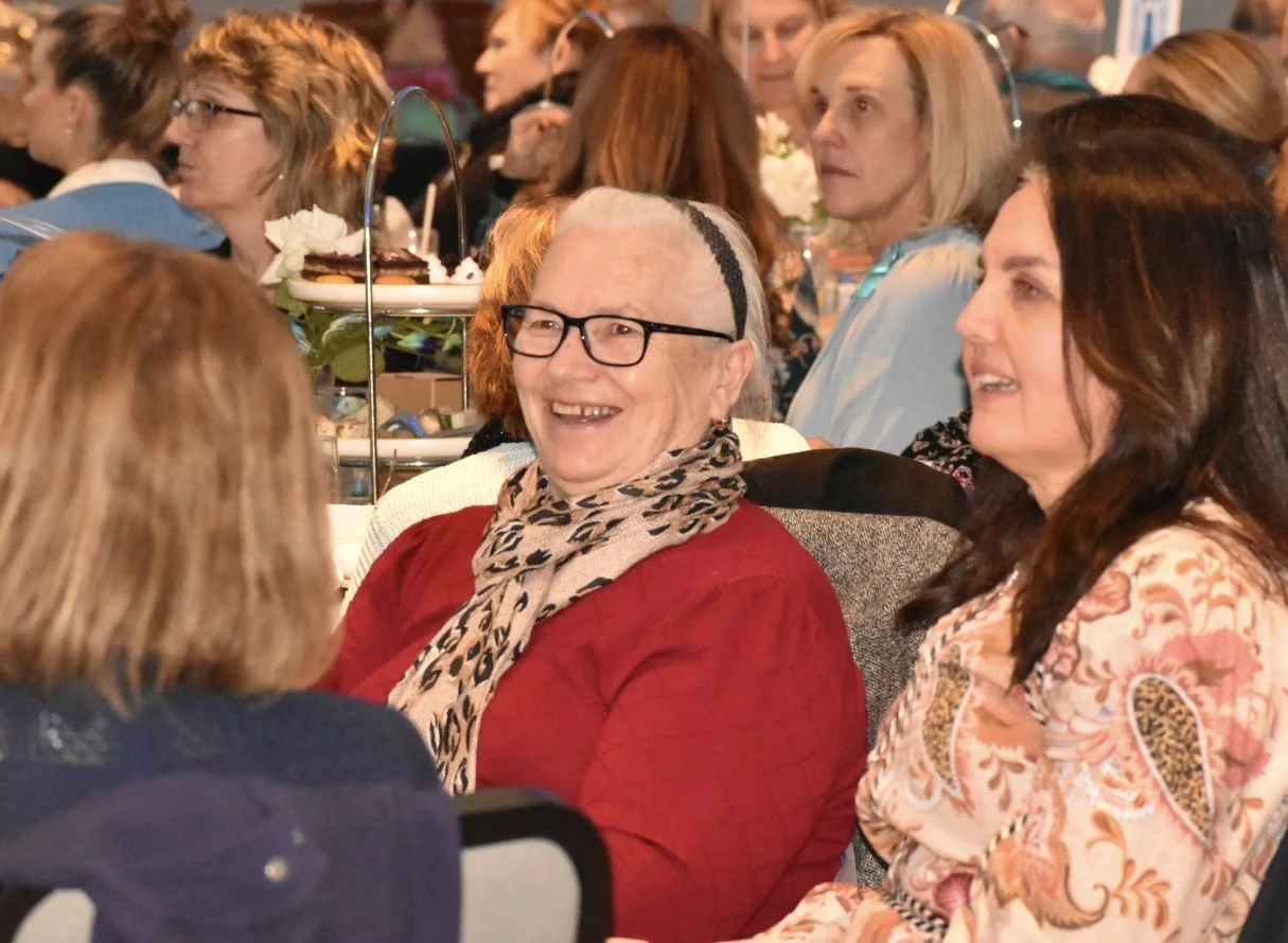 A group of women sitting at a formal event or gathering, some smiling and engaging in conversation, with decorative food stands in the background.