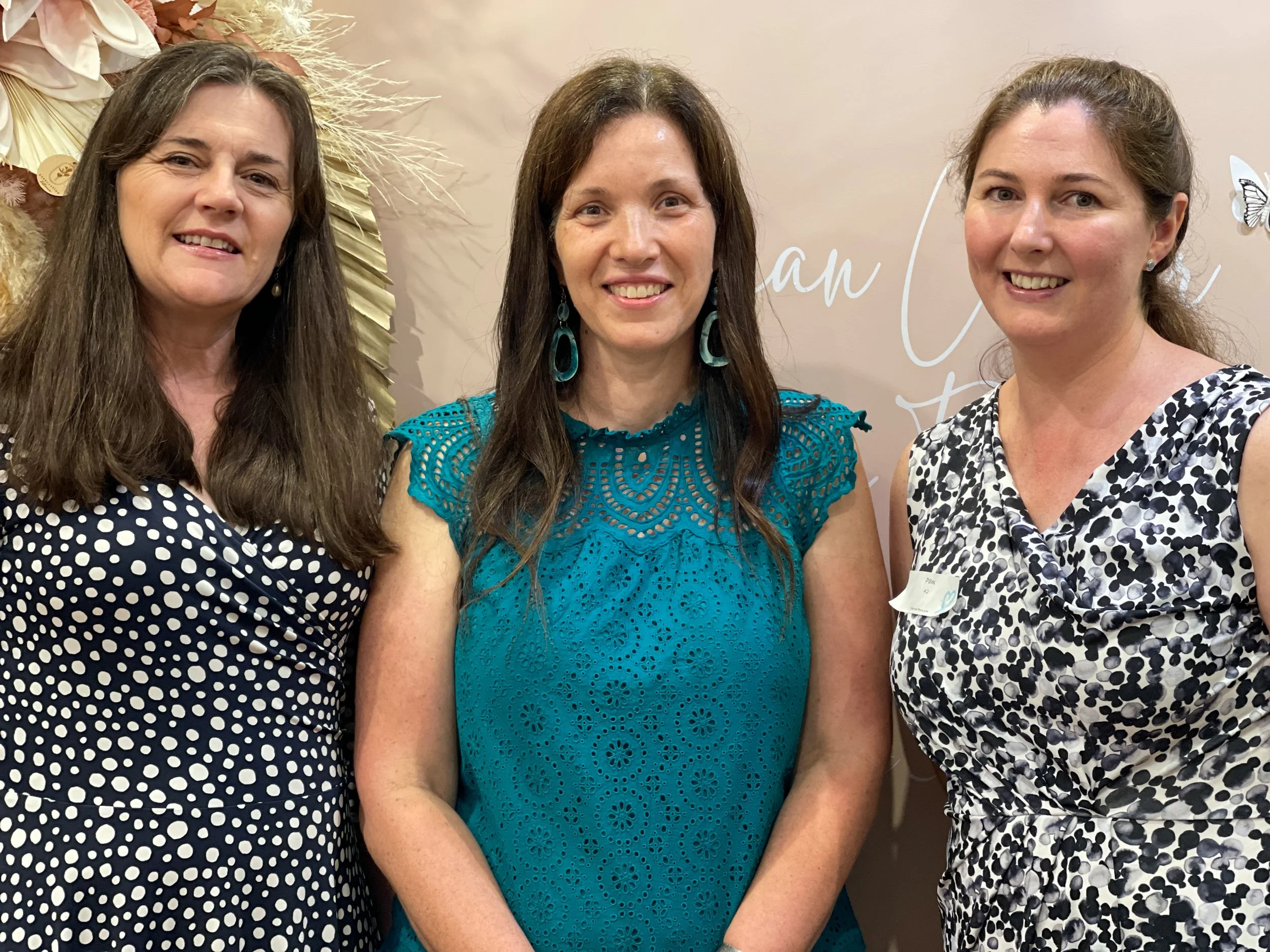 Three women standing together, smiling, at a social event in front of a beige backdrop with decorative elements and partially visible text.